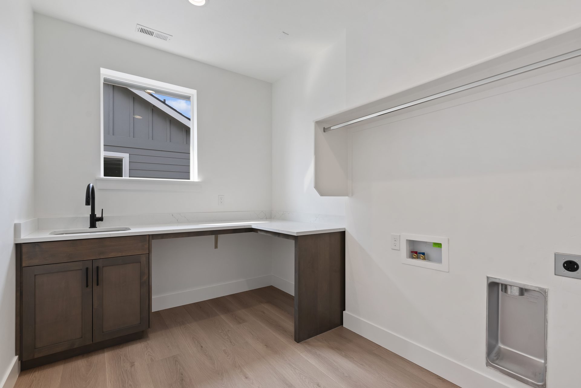 A room with a brown stained L shaped vanity with a single sink, a single window and a shelf with a single hanging rod and medium brown wood flooring.