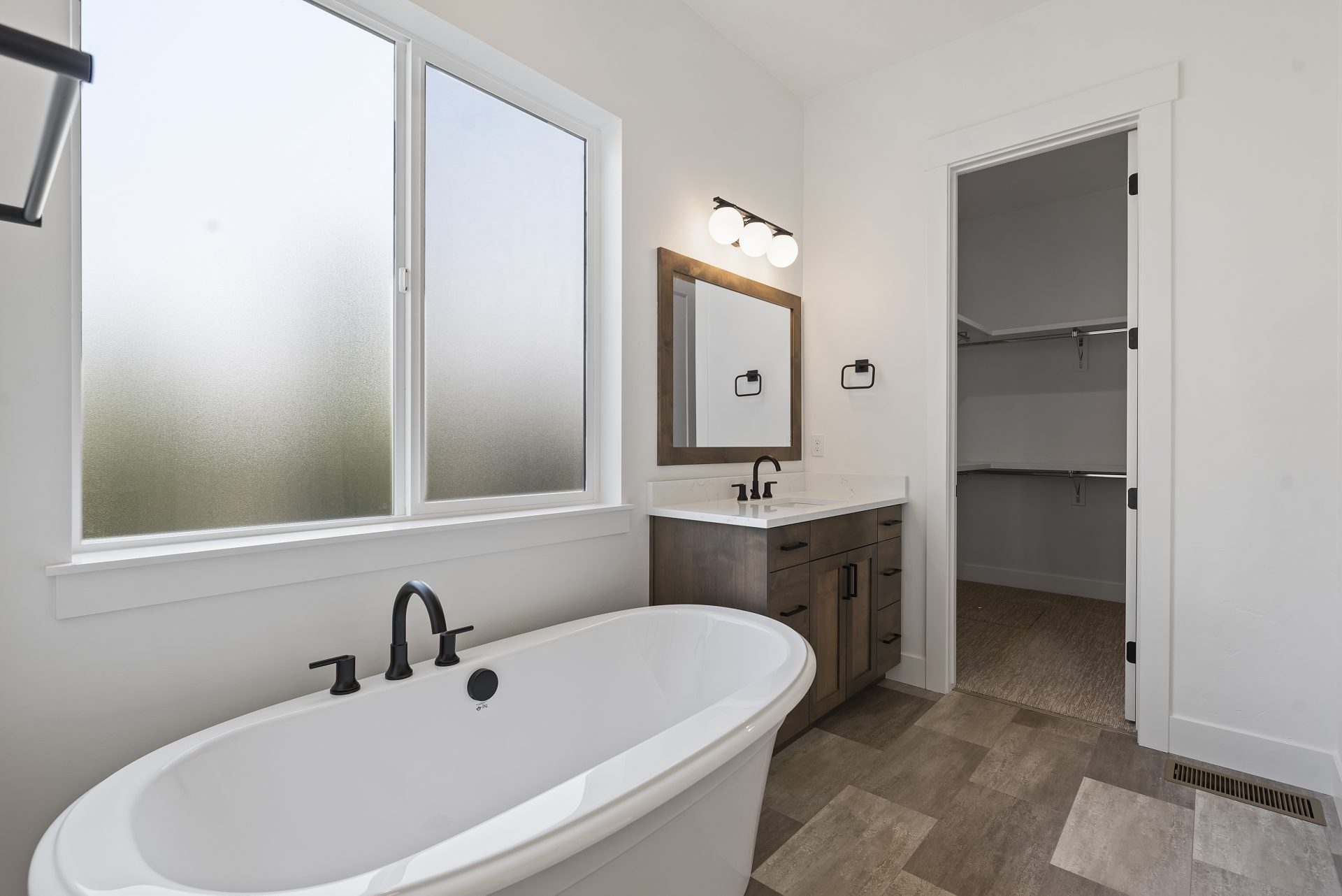 Bathroom with a free standing white tub with black finishes located underneath a large frosted window, a single vanity with black finishes, white countertop and brown stained wood, a mirror with matching brown stained frame a single light that includes 3 