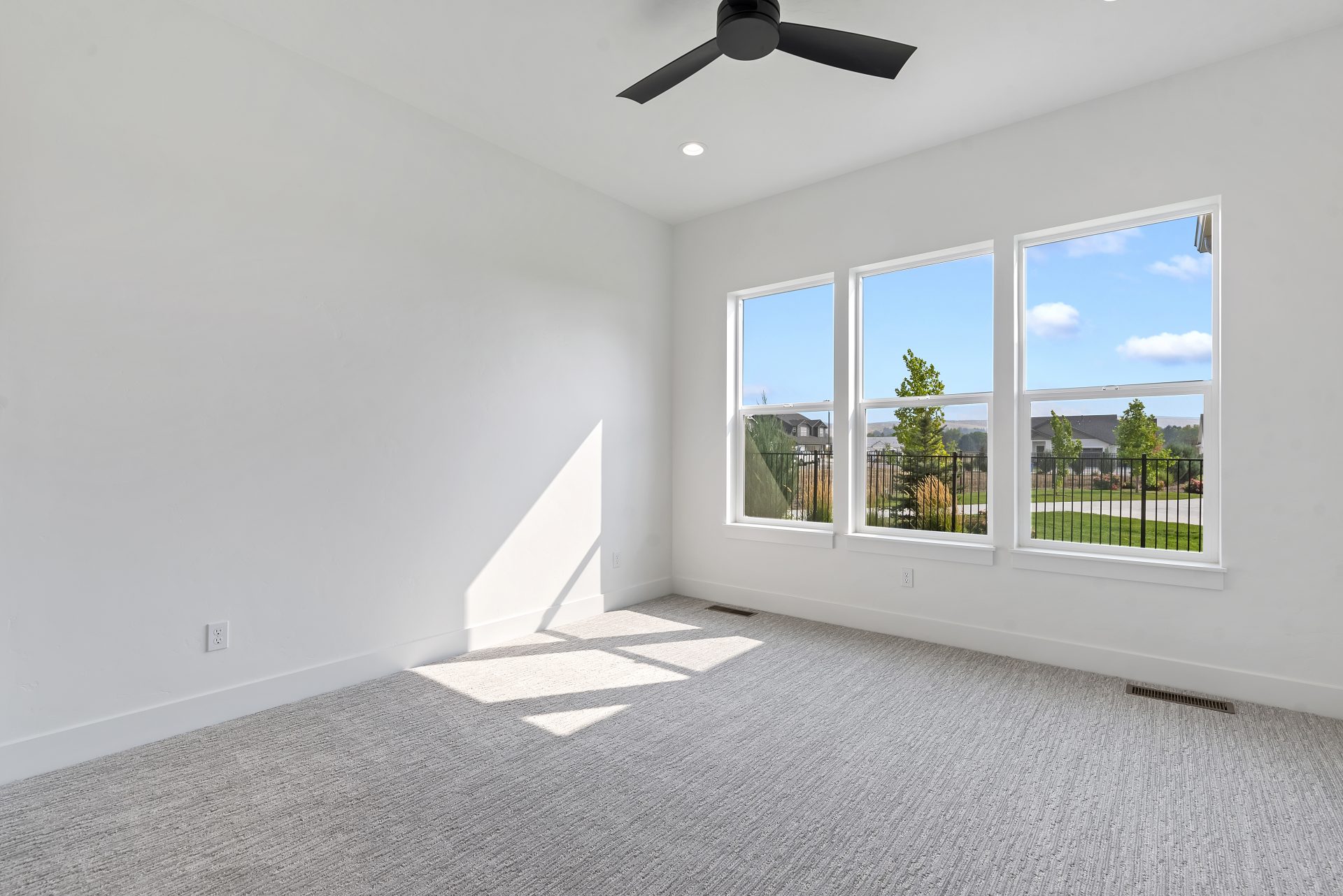 A bright room with light gray colored carpet, white walls, 3 windows appear to be 2'wide and 3' tall, and a dark colored ceiling fan.