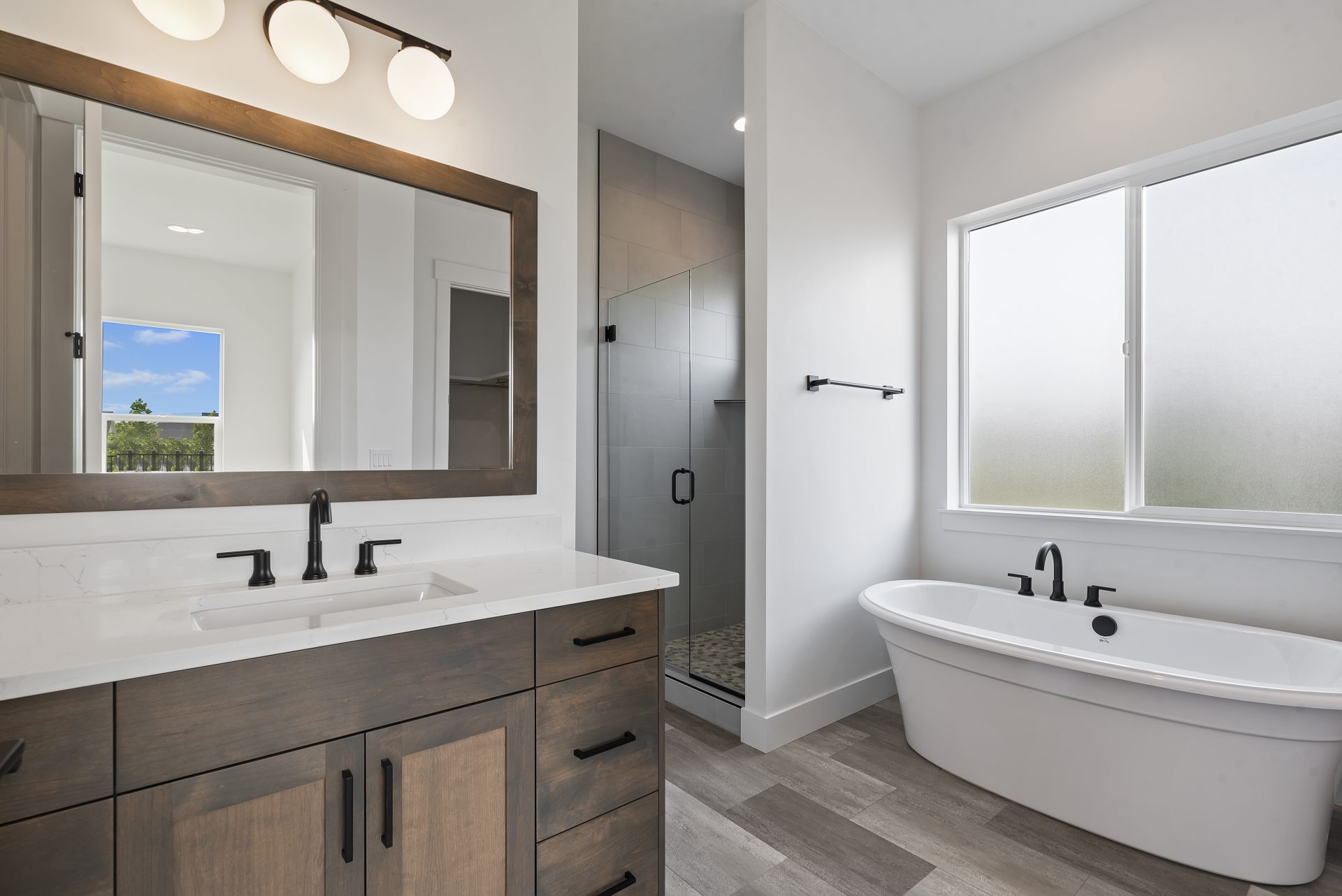 A bathroom with a single sink vanity, white countertop, black finishes, brown stained wood and a mirror that spans the length of the vanity with a wooden frame that matches the same stain color as the vanity. A fee standing tub sits below a large frosted 