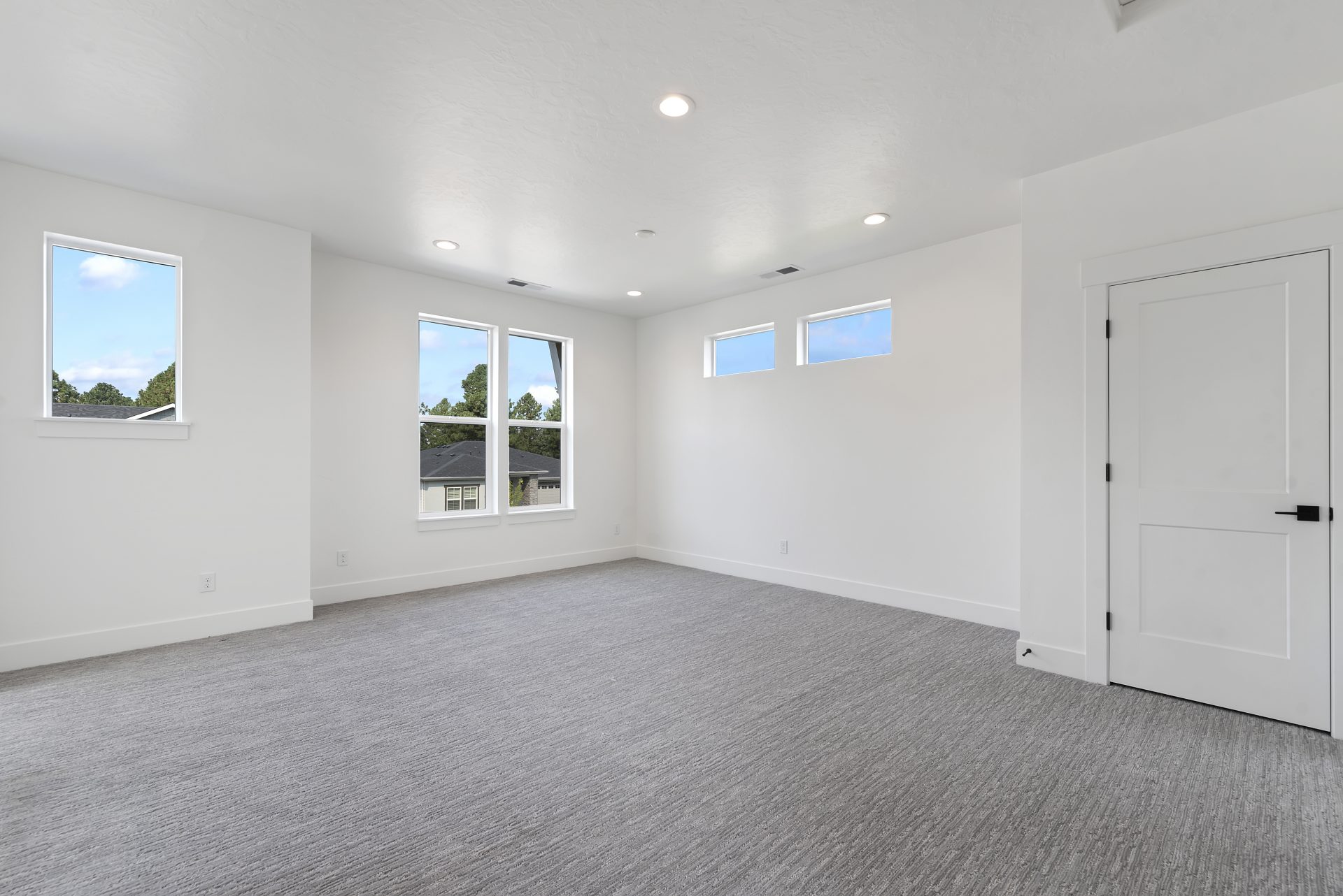 A bright bedroom with light gray carpet multiple windows and a single door.