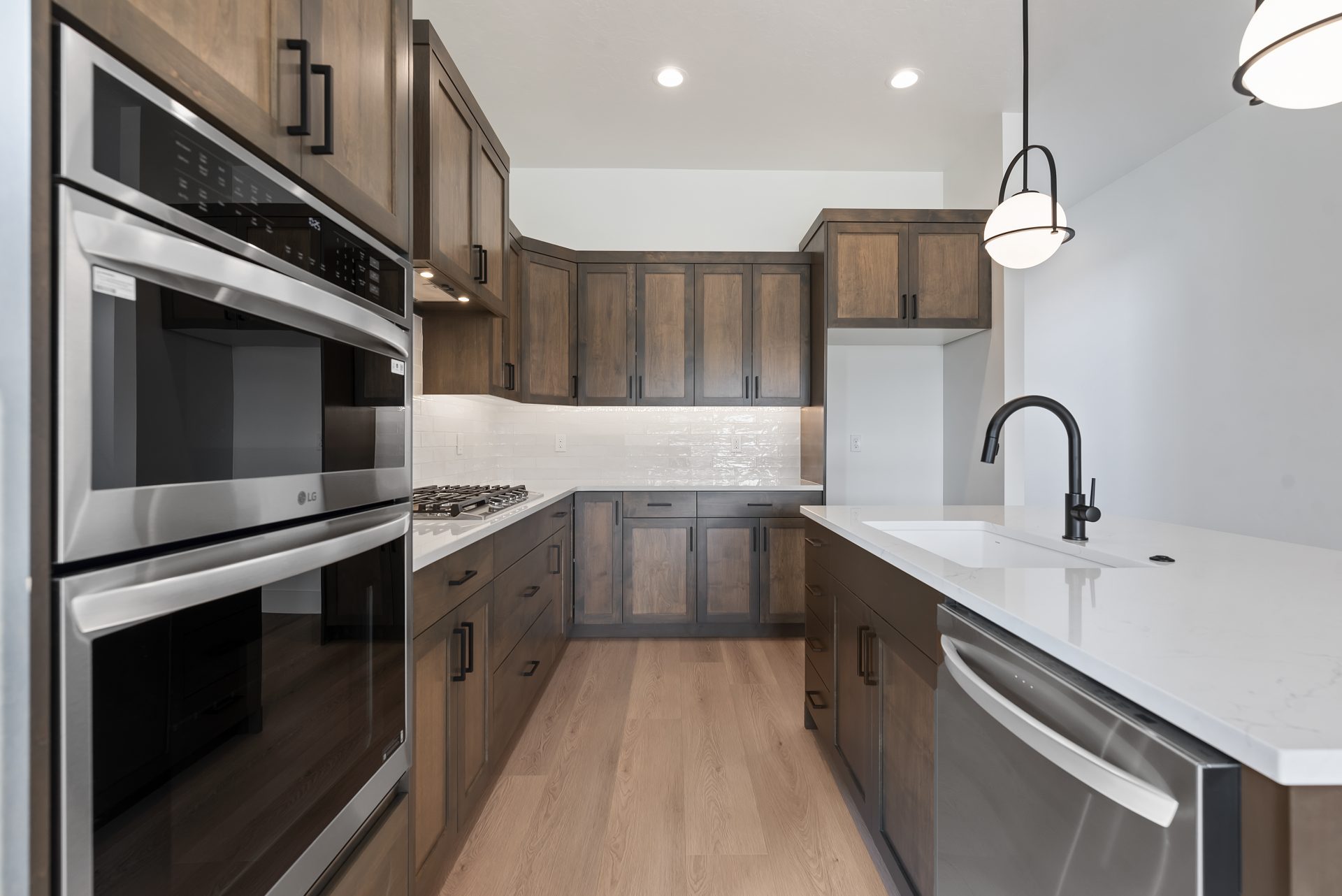 A kitchen area up close with view of the space between the island and the cabinetry along the wall with double ovens and a gas range, with lots of brown stained cabinetry, kitchen island with two hanging pendent lights, a sink with dark bronze fixture and