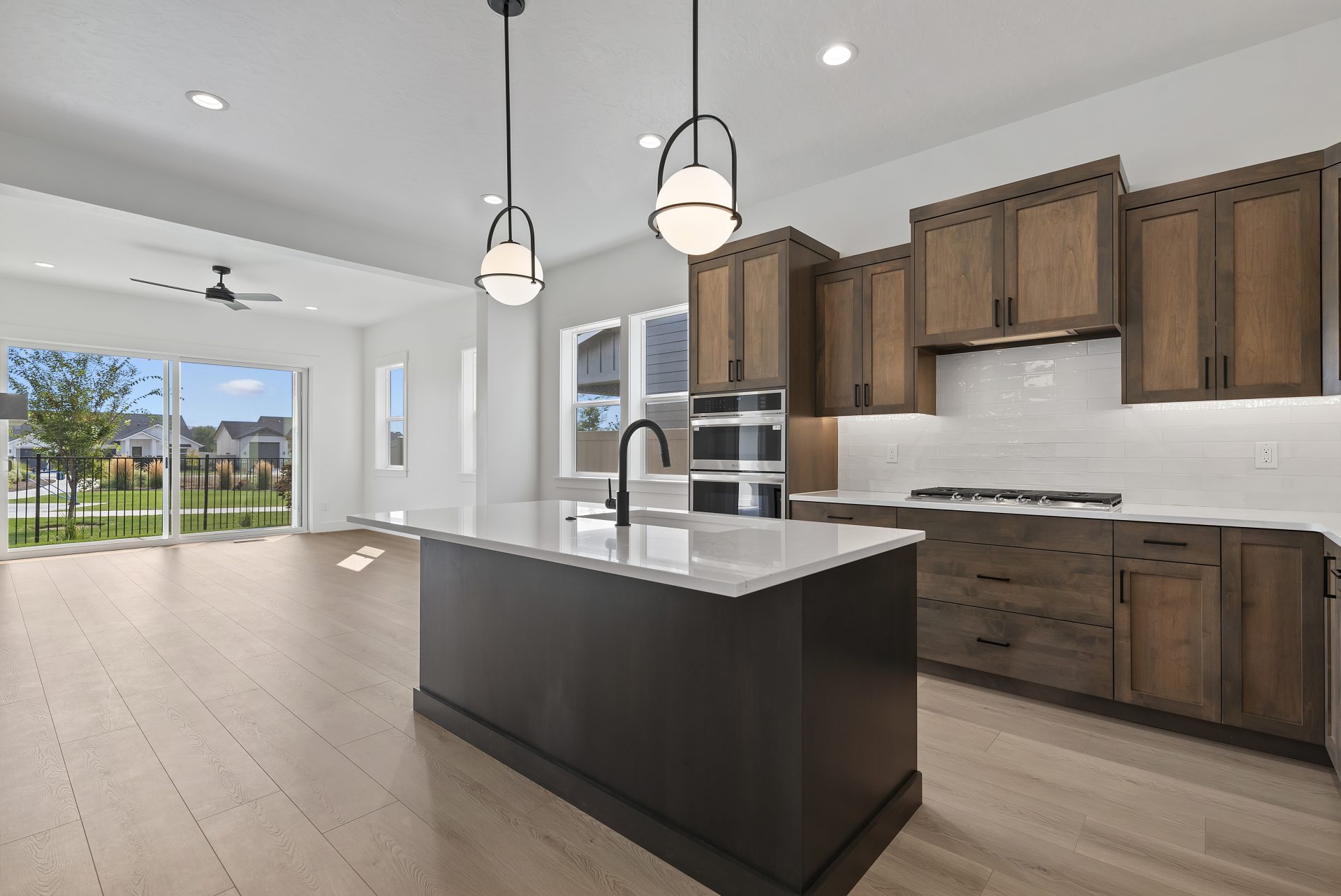 A view of the kitchen with a brown stained island with white countertop, a large dark bronze faucet, two pendent lights hanging above island, lots of brown cabinetry, white tile behind gas range and up the wall between white countertop and brown cabinetry