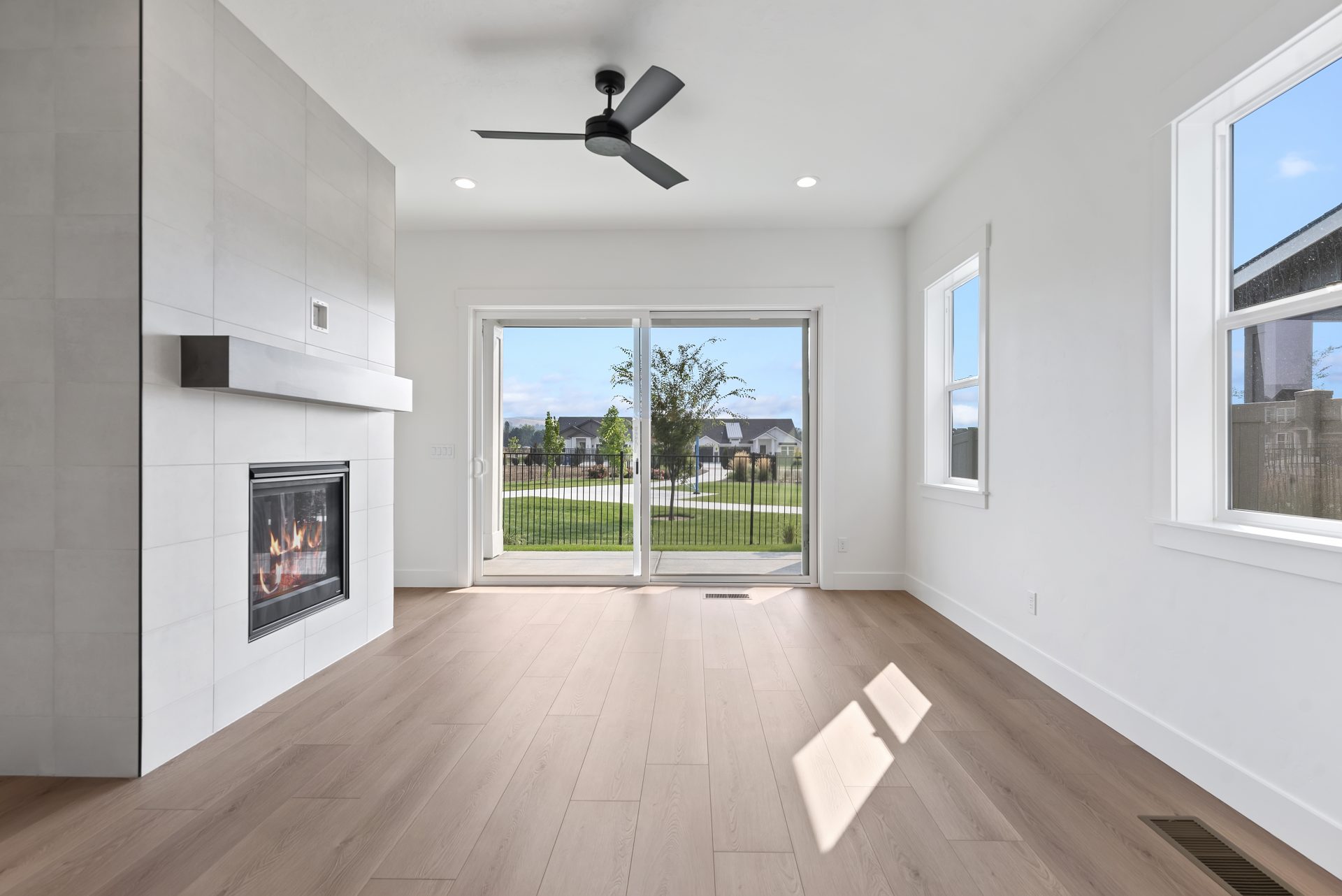 A bright room with medium brown wood flooring, a black ceiling fan, fireplace with gray tile floor to ceiling detail, a sliding glass door and two windows.