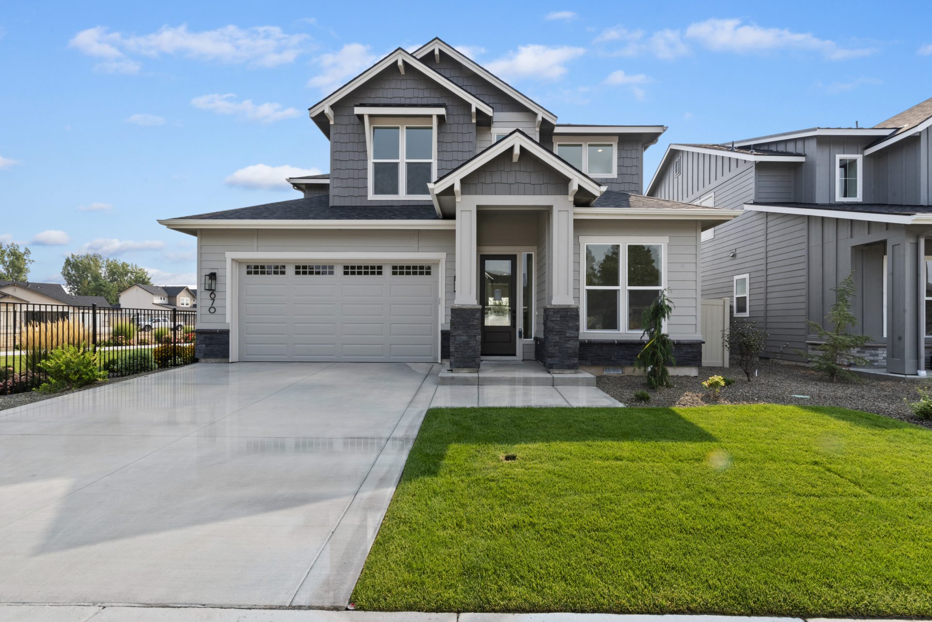 An exterior view of a modern two story home with gray shingles and a lighter gray of wood paneling, a covered porch and entry way with slate colored tile accent on the posts with a two car garage and green grass plus landscaping.
