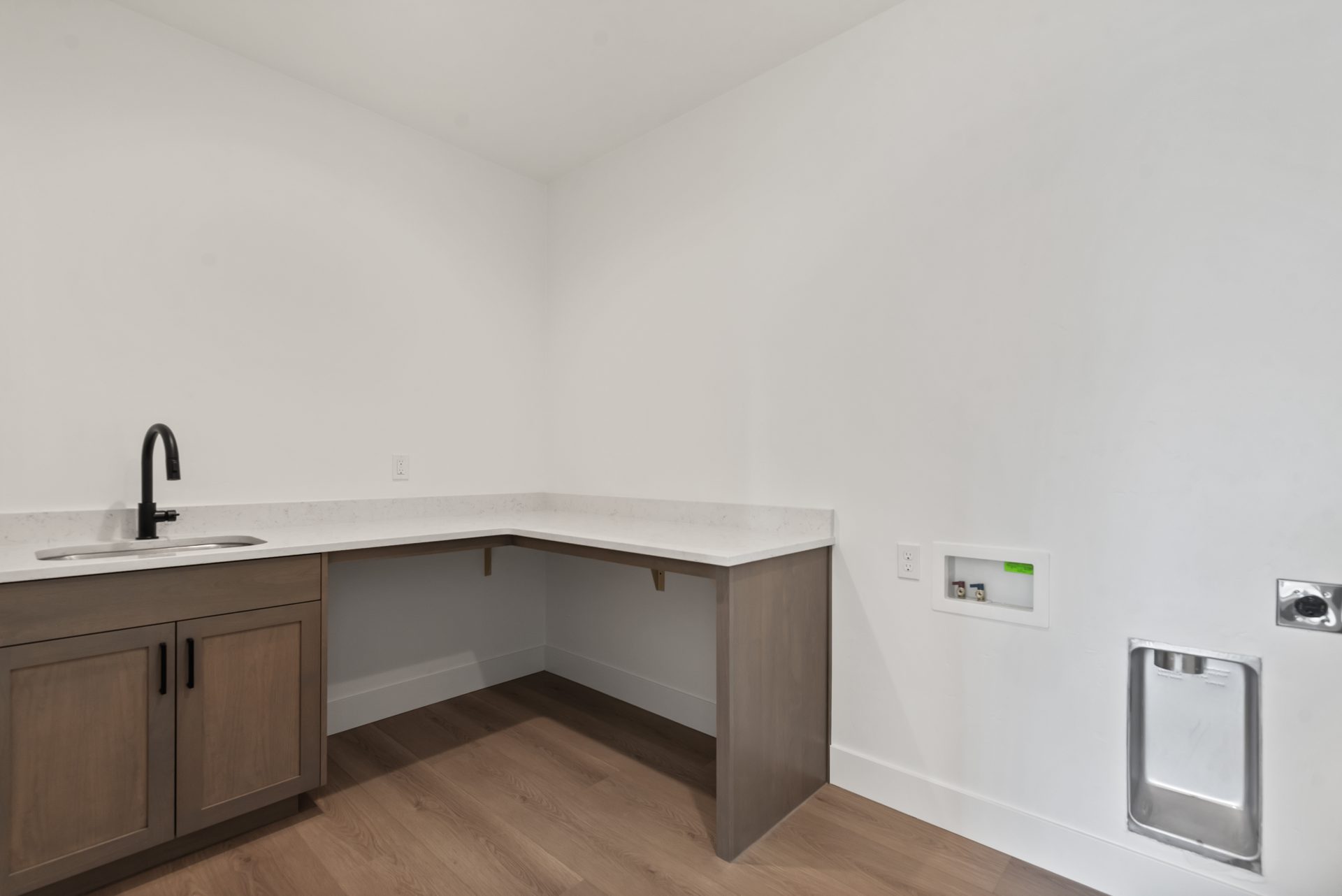 A laundry room with an L shaped vanity with a single sink, brown cabinetry and a white countertop with a short backsplash, white walls, washer and dryer not pictured.