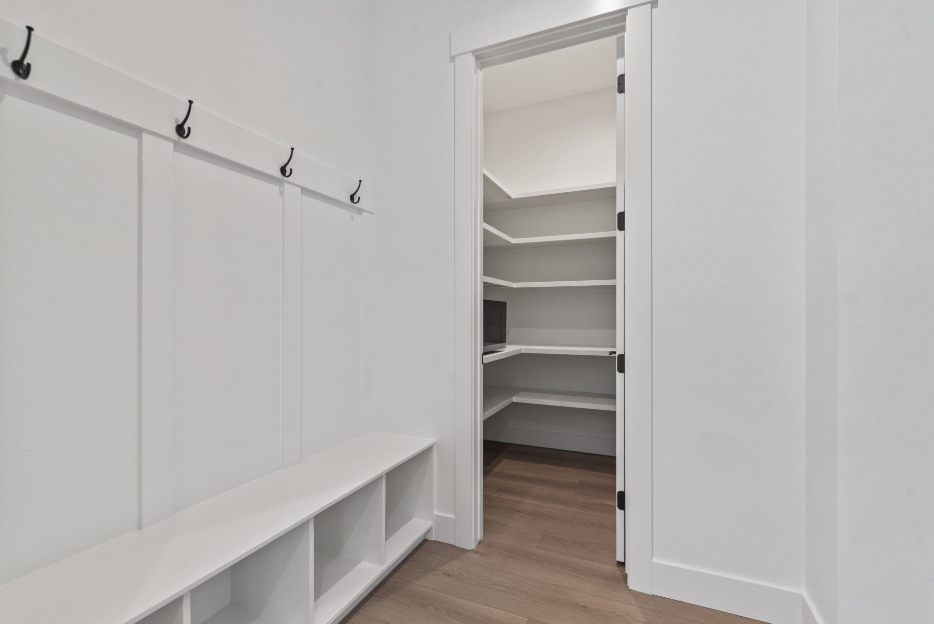 A room with white walls and white paneling with 4 hanging hooks, a bench with storage cubbies underneath, brown wood flooring and an open door showing storage with shelving.