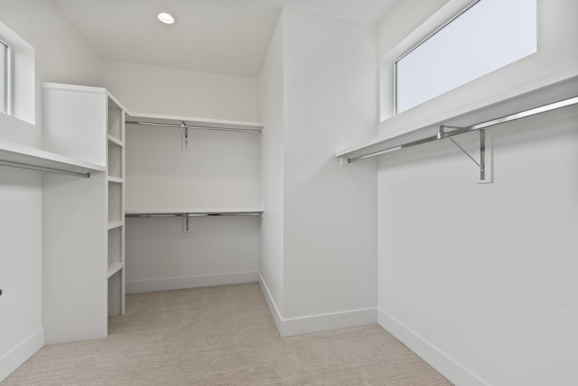 A large walk in closet with white walls and beige carpeting, with 4 steel hanging rods and multiple levels of shelving for storage.