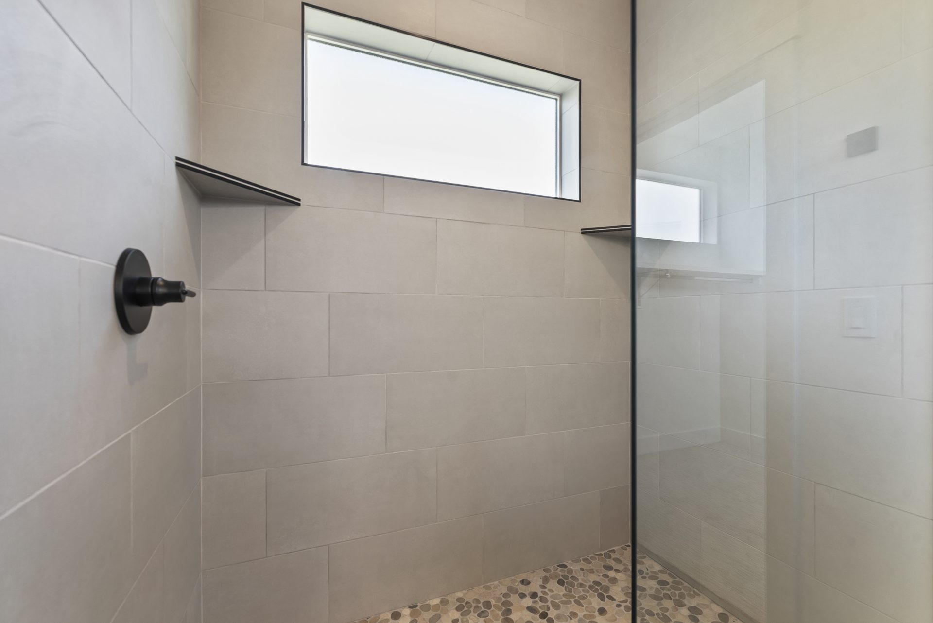 A large shower space with glass entry door, natural stone floor, wall to ceiling gray tile approximately 18" x 8"  with two shelves and black fixtures.