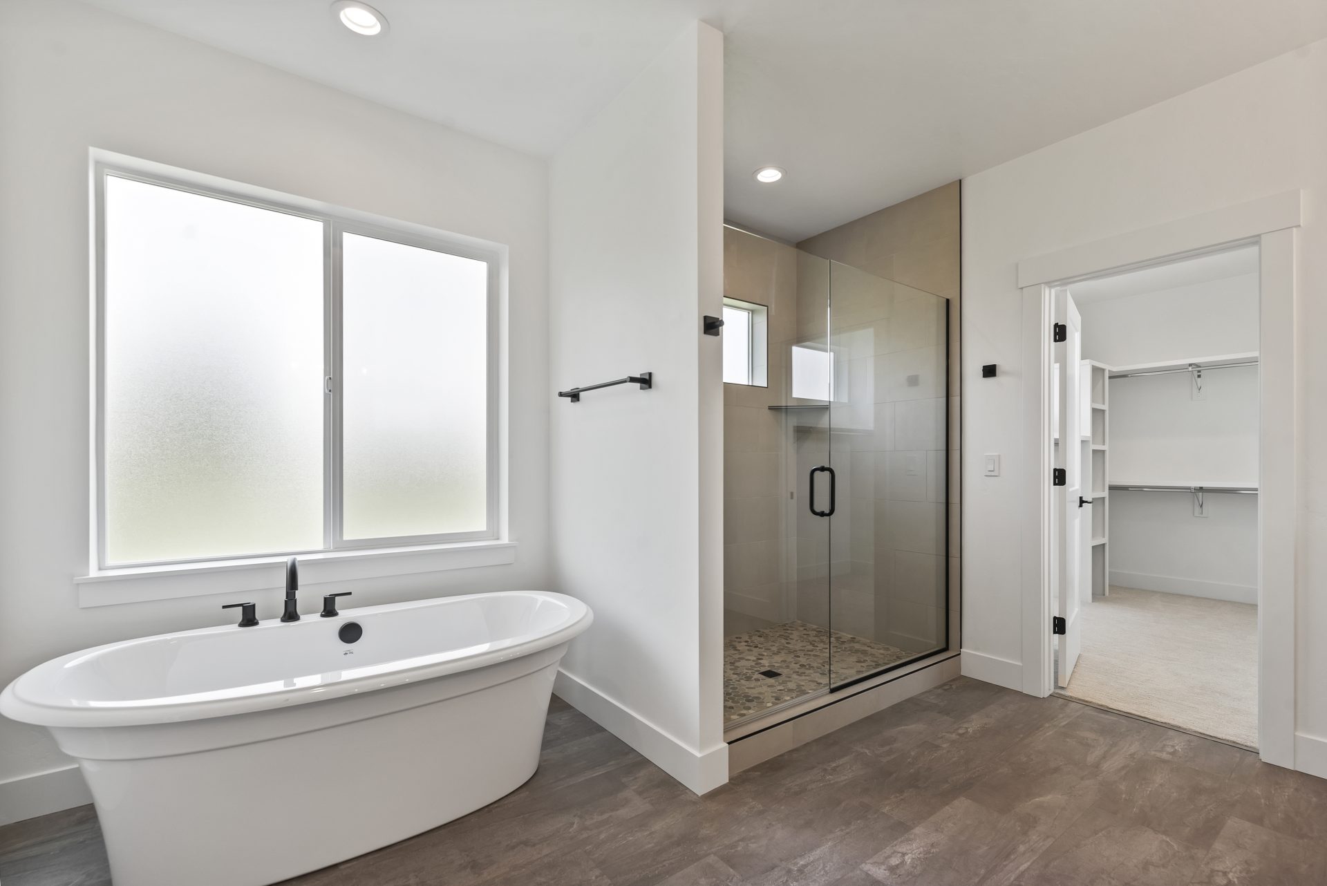 Bathroom with can lighting, a free standing tub with a large frosted window located above, a view of a large shower with glass door and black handle, a open doorway leading to a carpeted closet and large brown tile flooring.