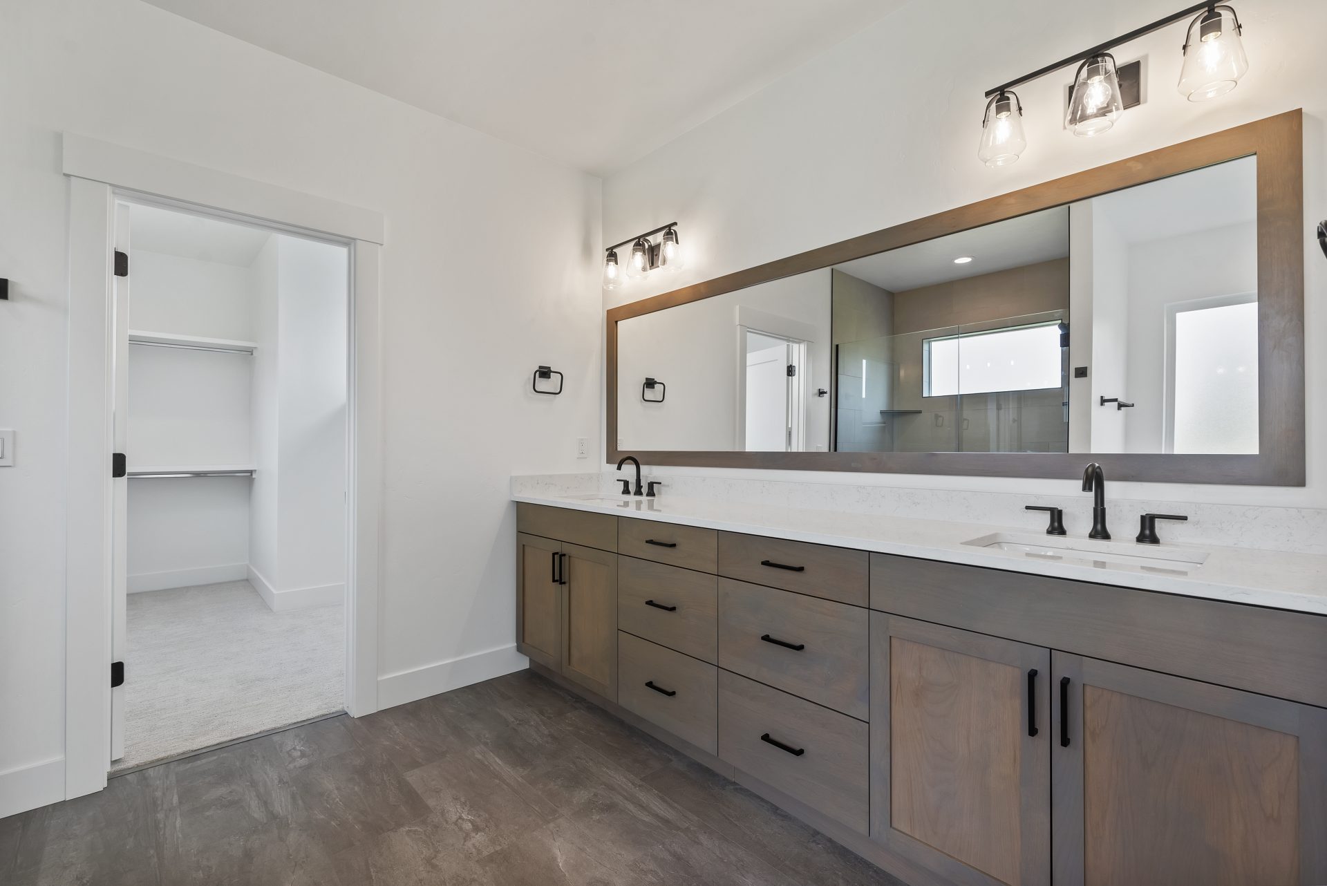 A bathroom with large brown tile flooring, a long vanity with brown stain, double sinks, white countertops, black fixtures, a wall mirror spanning the length of the vanity with a matching brown trim, two light fixtures located above the mirror, each havin