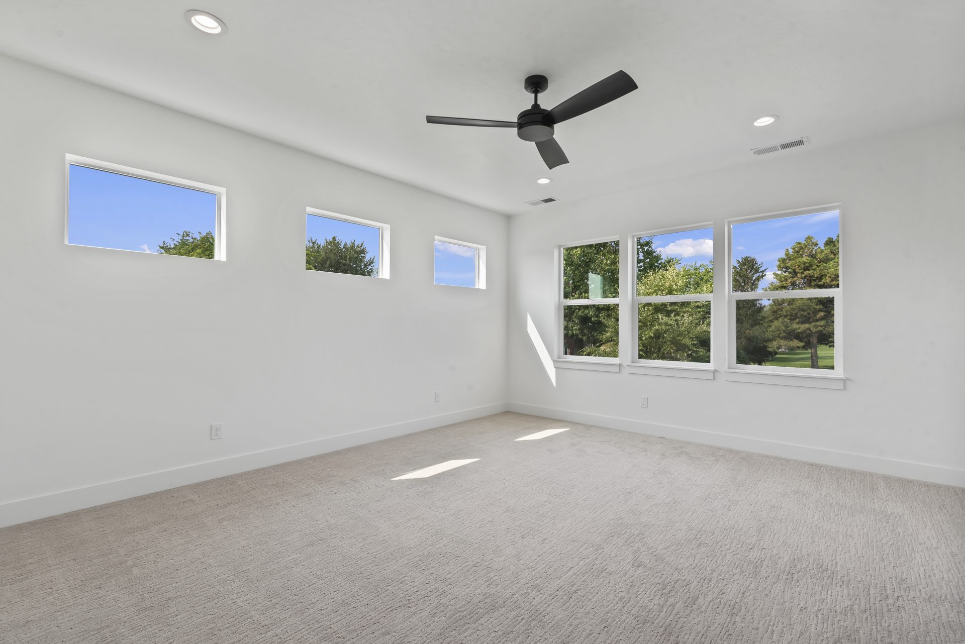 A bedroom with 6 windows, 3 are 2 feet wide by 2 feet high on left wall, and the other 3 are 2 feet wide by 3 feet high on right wall, light gray carpet, white walls and a black ceiling fan.