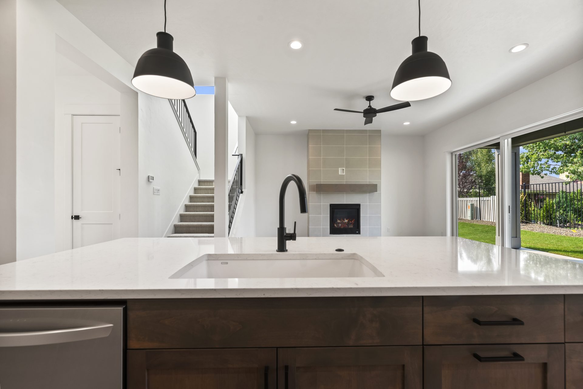 A kitchen island up close with a sink with black faucet, a dishwasher, brown stained cabinets, two black hanging pendent lights, a view of a staircase, a pillar and a fireplace with floor to ceiling gray tile detail and a brown stained wooden mantle, and 