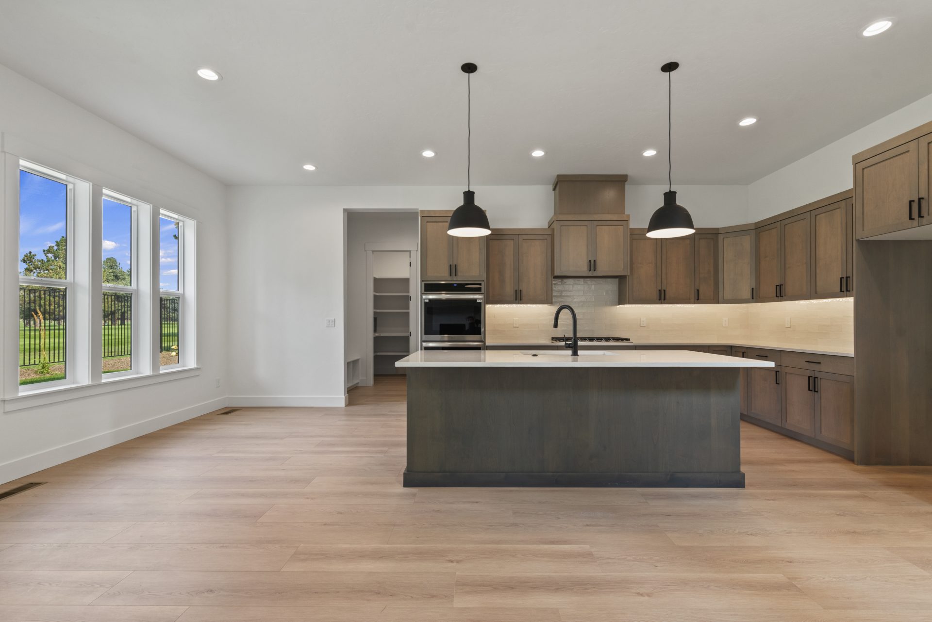 A view of the kitchen complete with a kitchen island stained with a dark brown color, white countertop, kitchen sink and a black faucet. There is a ton of cabinetry in a medium brown stain color, with long countertop space, a gas range with countertop to 