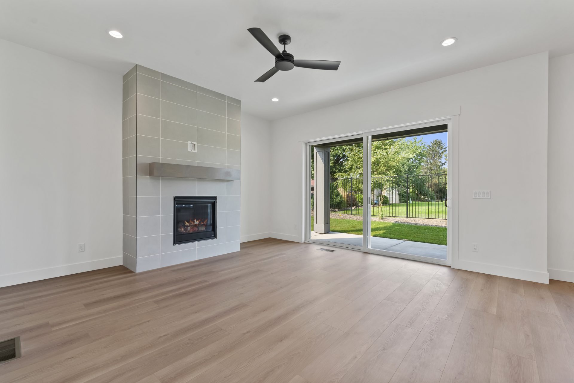 A bright living room with brown wood flooring, a sliding glass door, a fireplace with gray tile from the floor to the ceiling, can lights throughout the space and a black ceiling fan.