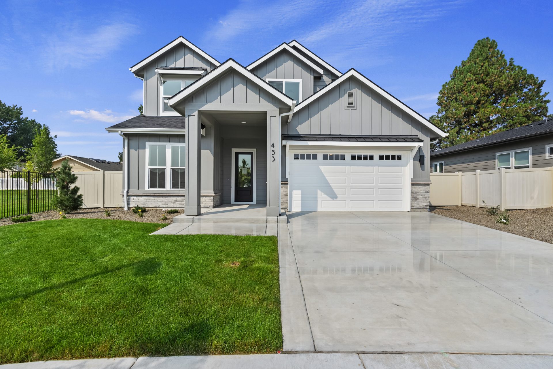 A modern home with gray siding, white two car garage door, covered patio and entry way leading to a front door with glass detail. A landscaped front yard with grass, rock, trees and bushes.