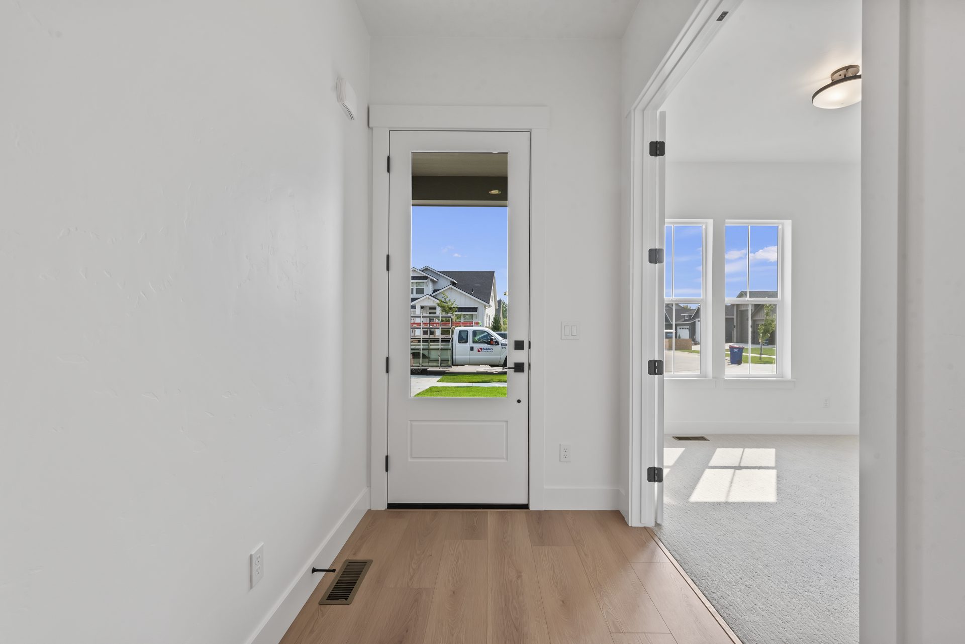 A entry way with a door with glass detail, white walls, soft brown wood flooring and a large opening to another room on the right.