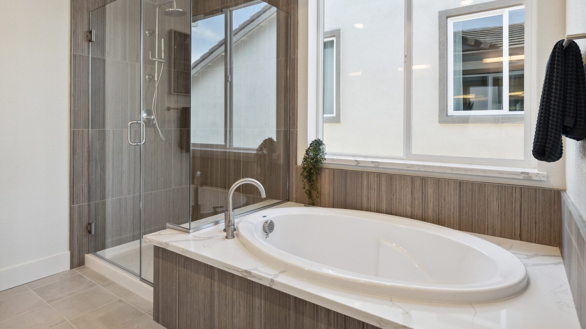 A bathroom with a large soaking tub with brown tile surrounding it. Above the tub is a large window, and behind the tub is a stand in shower encased in a glass with a chrome knobs and fixtures.