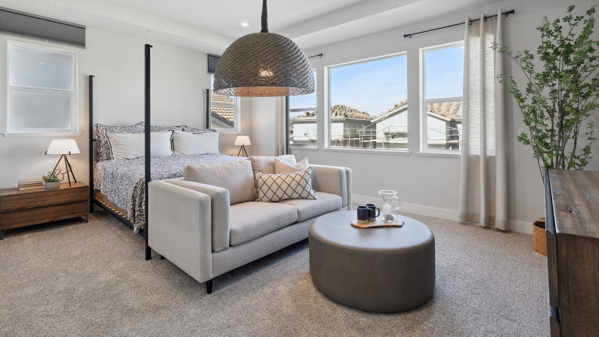 A large bedroom furnished with a bed and small sofa, nightstands and dresser, with gray carpet and large windows on two adjacent walls with window coverings.