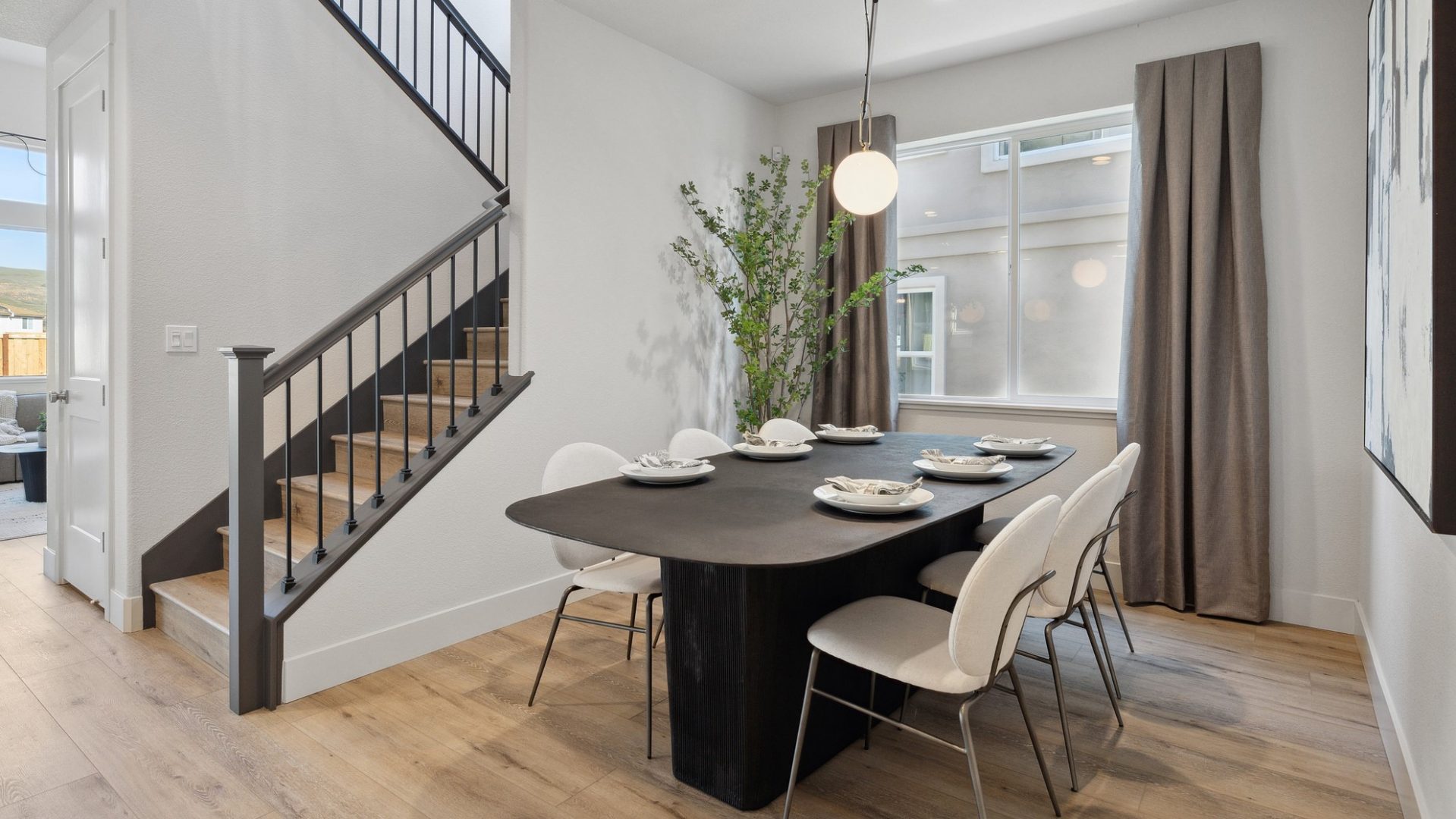 A dining nook with a dark colored dining table and six chairs with a window with long window covers, a pendant light hanging from the ceiling and a staircase to the left.