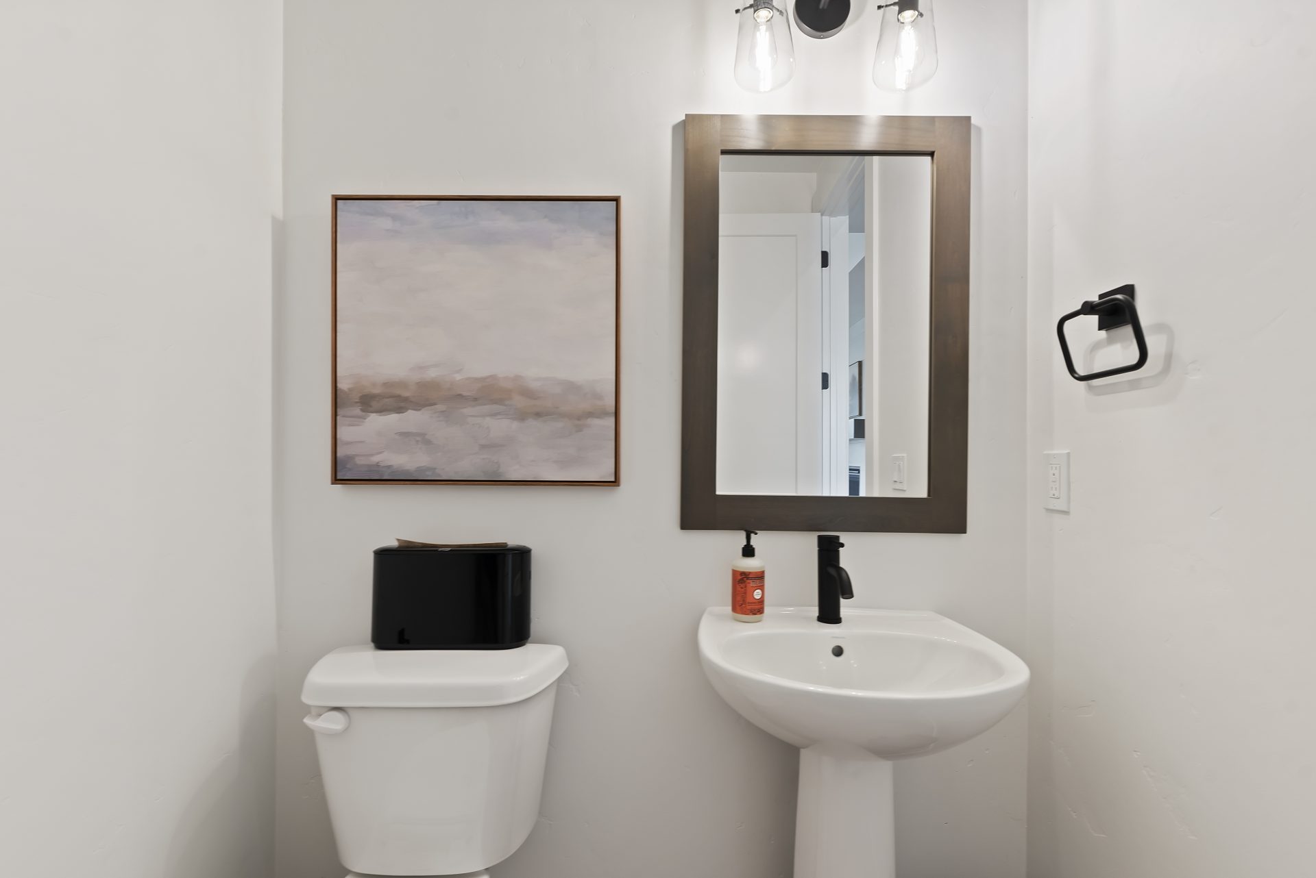 A powder room with a toilet, and white porcelain pedestal sink. with a framed mirror and light fixture located above.