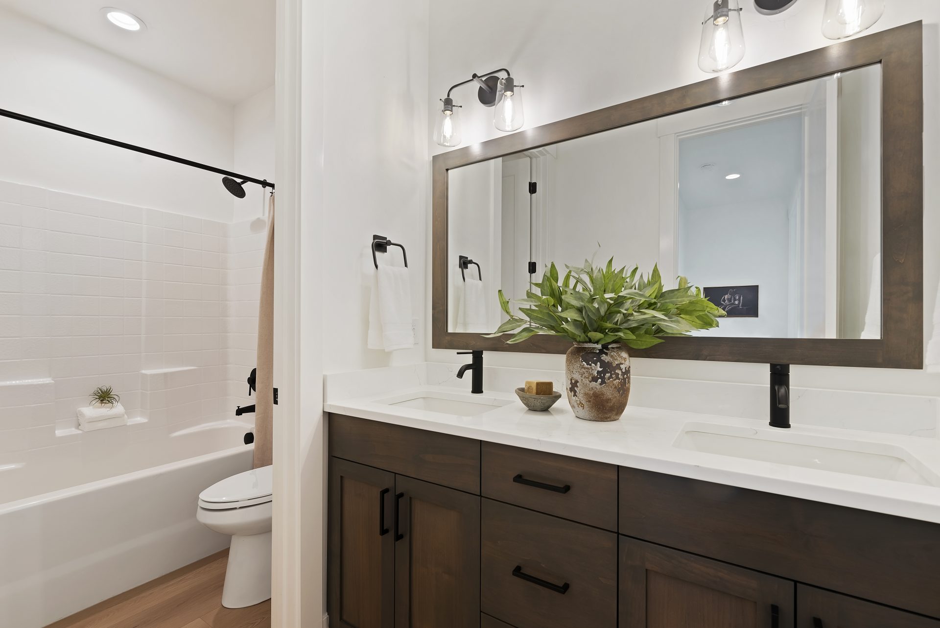 Bathroom with a dual sink vanity, with brown stain grade cabinets, black pulls and faucets, white countertop with 6" inch backsplash, a framed mirror spanning the length of the vanity and two light fixtures above. To the left a tub/shower combo and toilet
