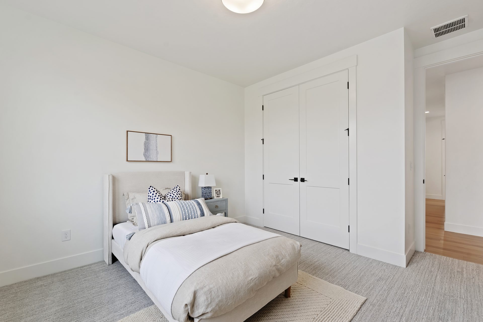 A bedroom staged with a twin bed and furniture items, gray carpet light color walls,  and a light fixture in the ceiling.
