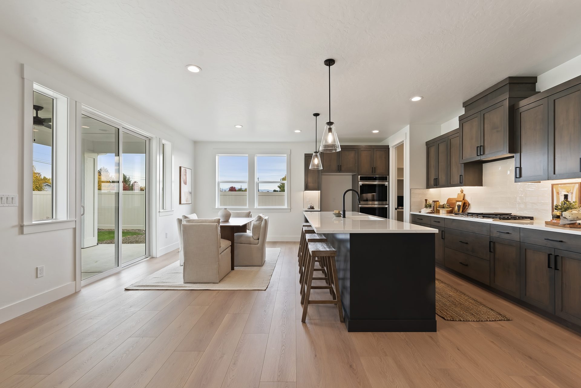 A staged dining area with a table and 4 chains, a kitchen complete with a kitchen island with four seats,  upper and lower cabinets with stainless steel appliances and white subway tile from countertops to cabinets.