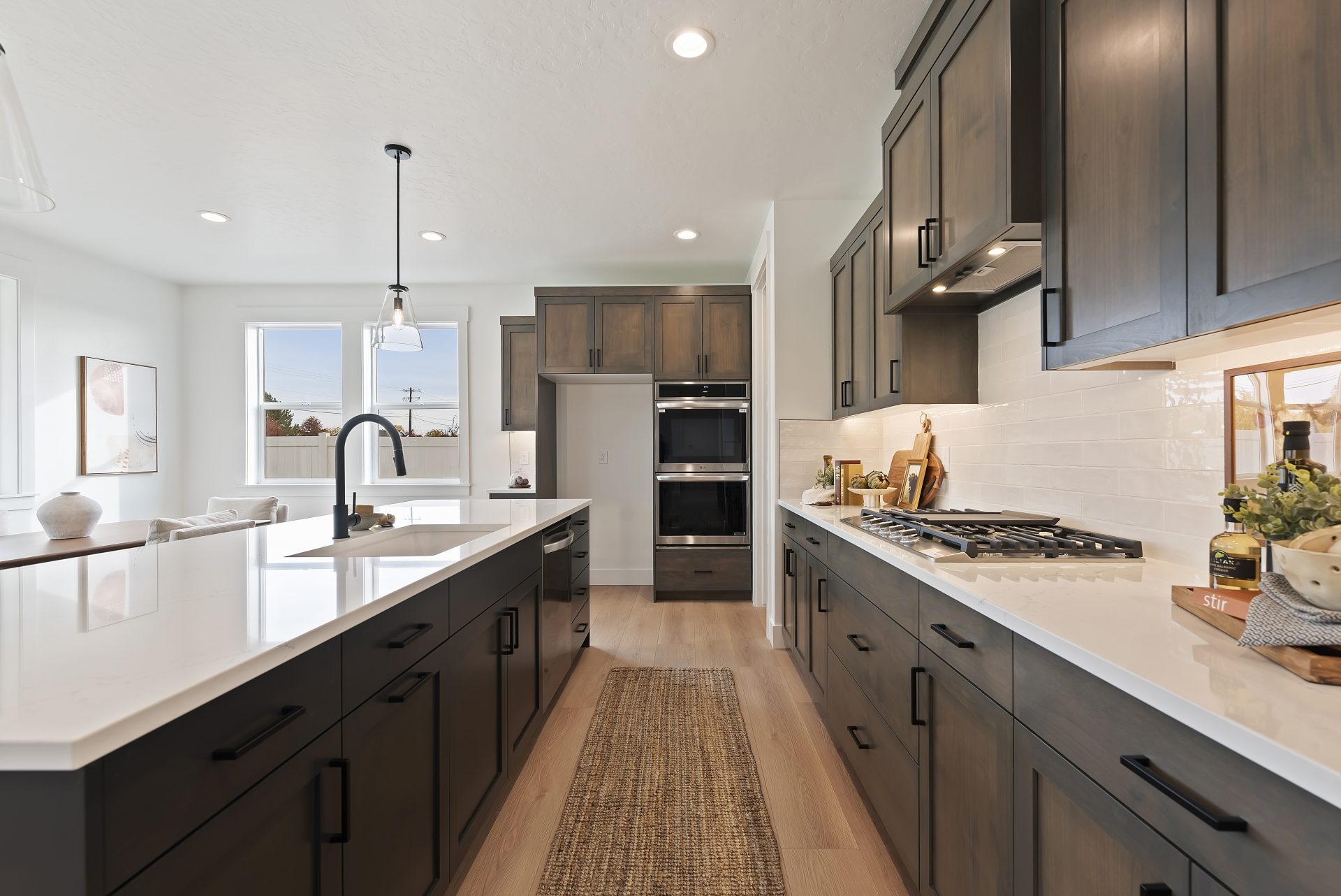 A kitchen area with a large kitchen island, brown stain grade cabinets with black pulls, white color countertops and a undermount sink with black faucet and a stainless steel dishwasher. On the opposite side of the island are the upper and lower cabinets
