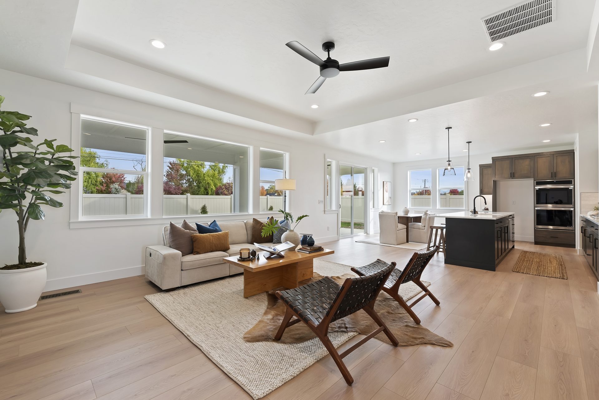 A staged living room with a couch, chairs and coffee table. Light brown wood floors, a gas fireplace with floor to ceiling tile and a mantle, can LED lighting in the ceiling and a ceiling fan over the seating area. Windows span across the back wall.