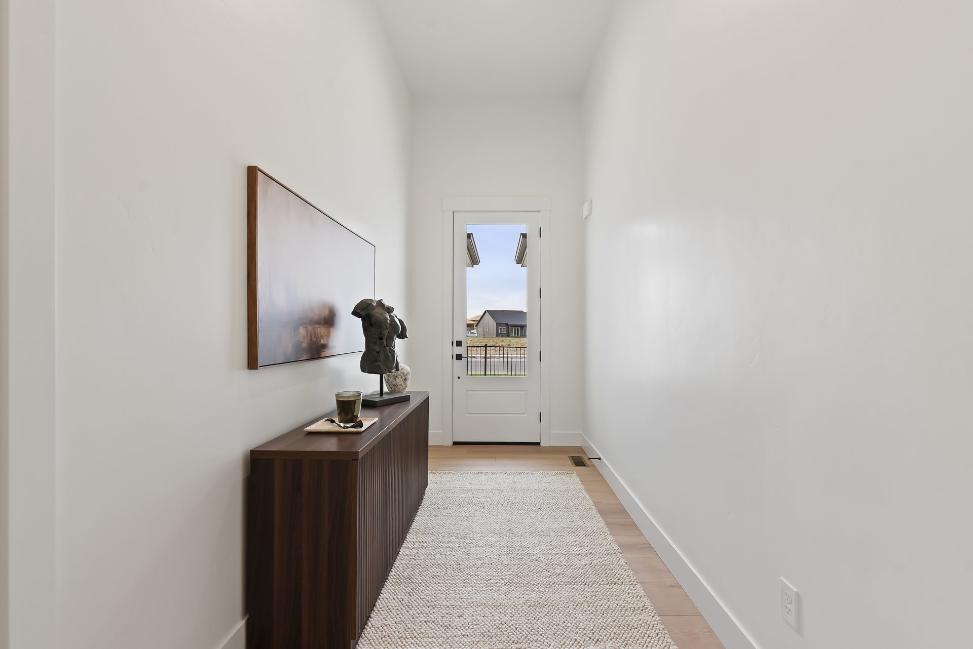 Interior entry way with a glass panel door, light brown wood floor with a runner rug and a credenza with various decor items and a hanging wall art located above.