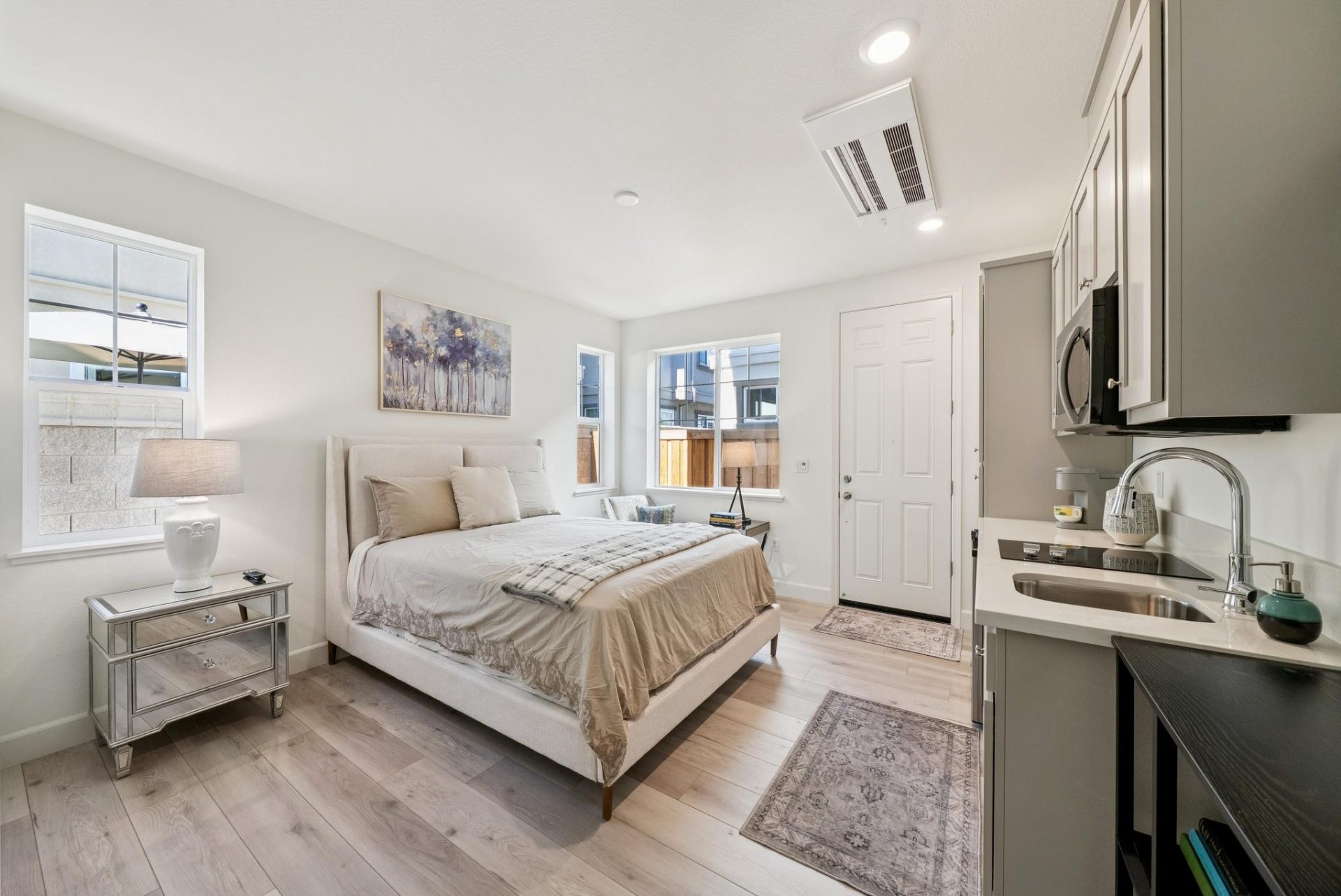 A furnished room with a bed and nightstand with lamp. To the right is a kitchen area with a single wall of cabinets, sink, and cook top with microwave.