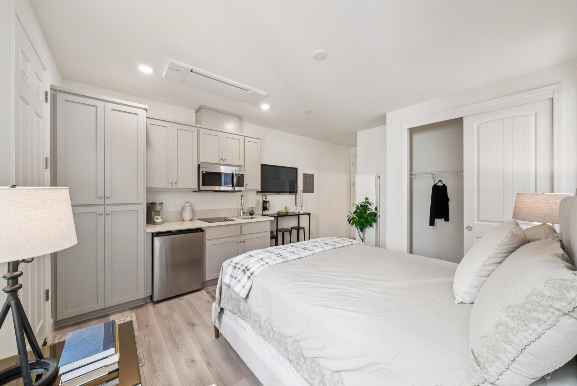 A furnished room equipped with a kitchenette including cabinetry, dishwasher, microwave, sink and cooktop. Furnishings include a bed, and nightstand with lamp. To the right is a closet space, the flooring is of light wood.