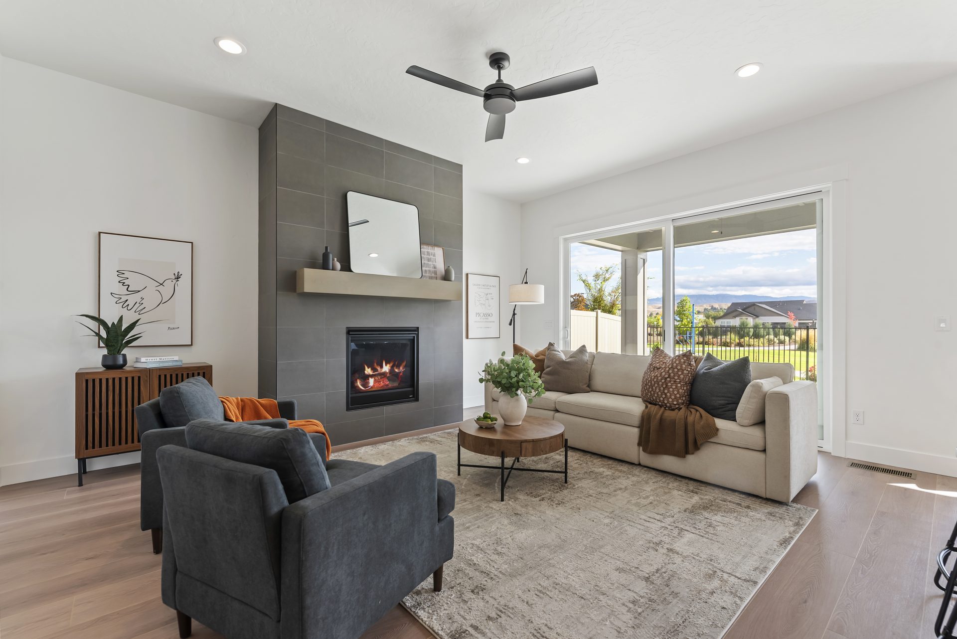 A living area furnished with a cream color couch and two dark blue lounge chairs, an area rug, round table and other décor items. There is a lit fireplace with floor to ceiling tile detail.