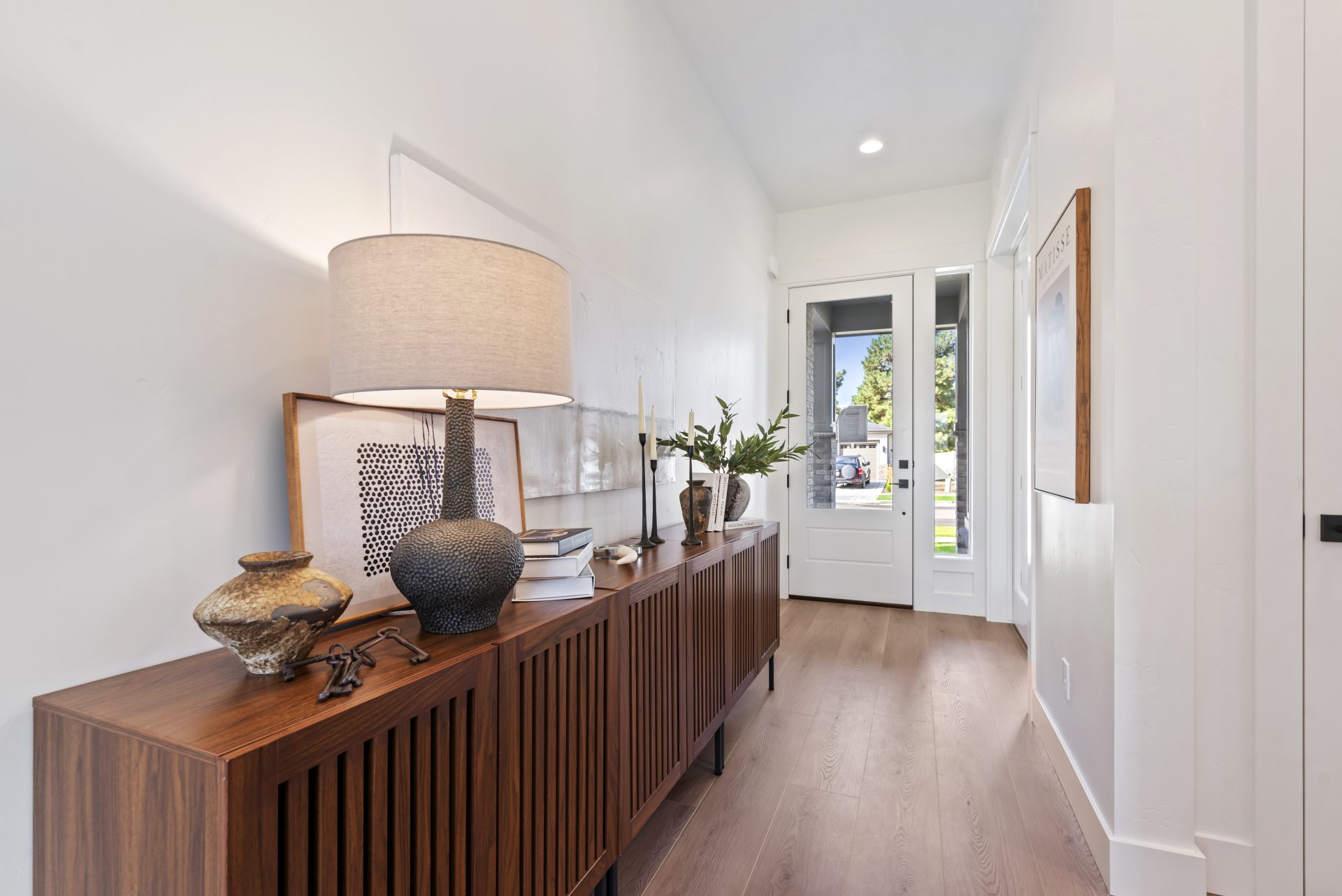 An entry to a home with a long credenza with various décor items on it, medium brown wood flooring and a front entry door white color with a glass panel.