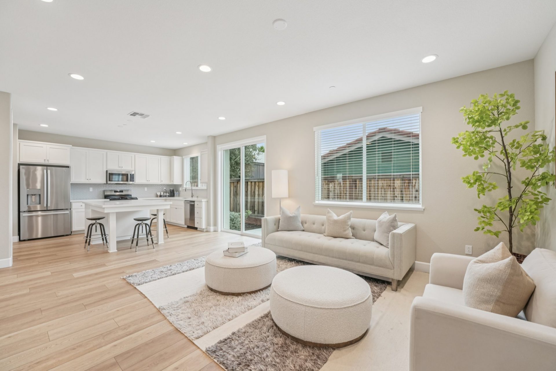 A staged living room with a seating area with light colored couches and chairs, an area rug, and two round ottomans. The walls are a light beige with white baseboards and light colored wood flooring.