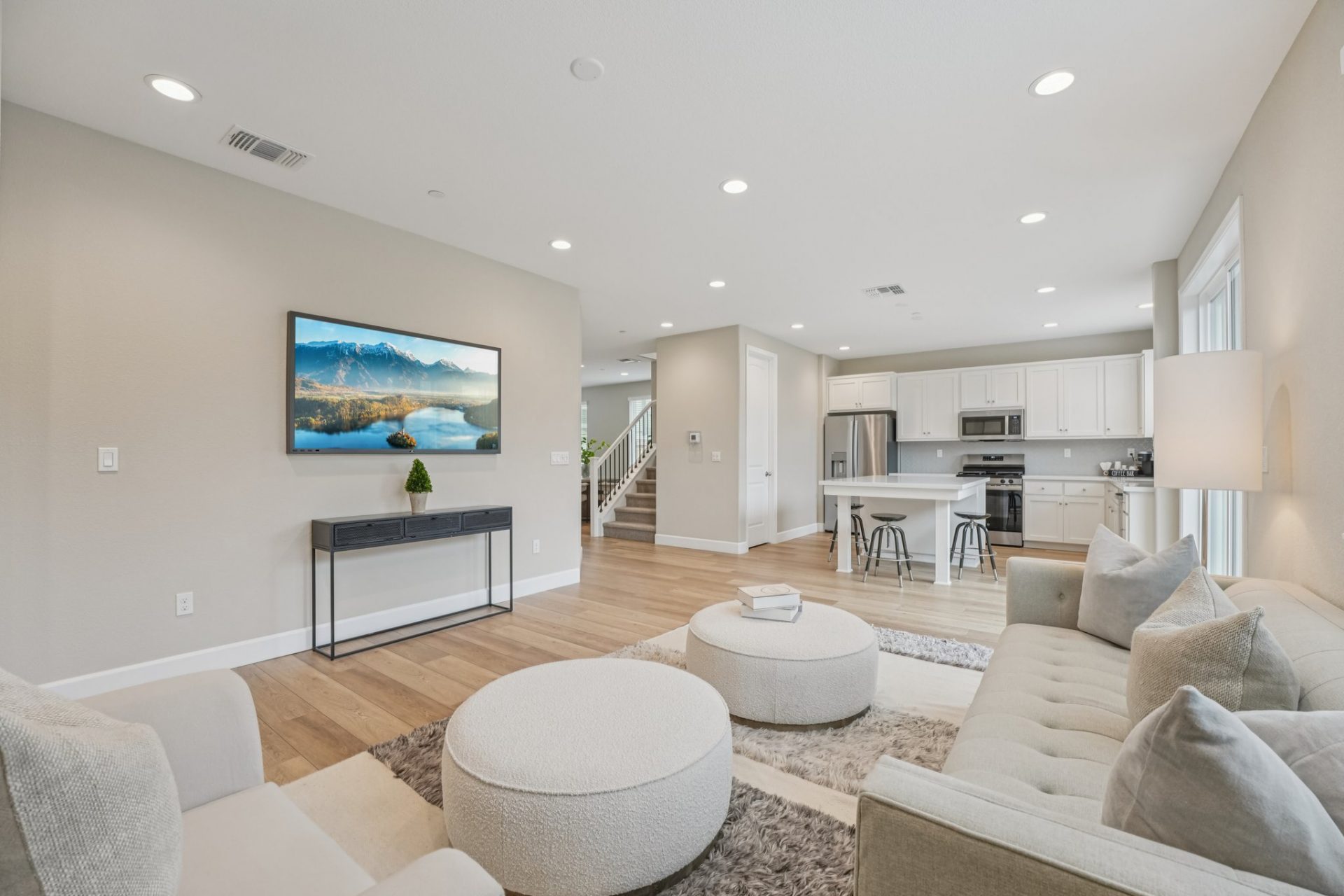 A staged living room with a seating area with light colored couches and chairs, an area rug, and two round ottomans. The walls are a light beige with white baseboards and light colored wood flooring and there is light décor pieces.