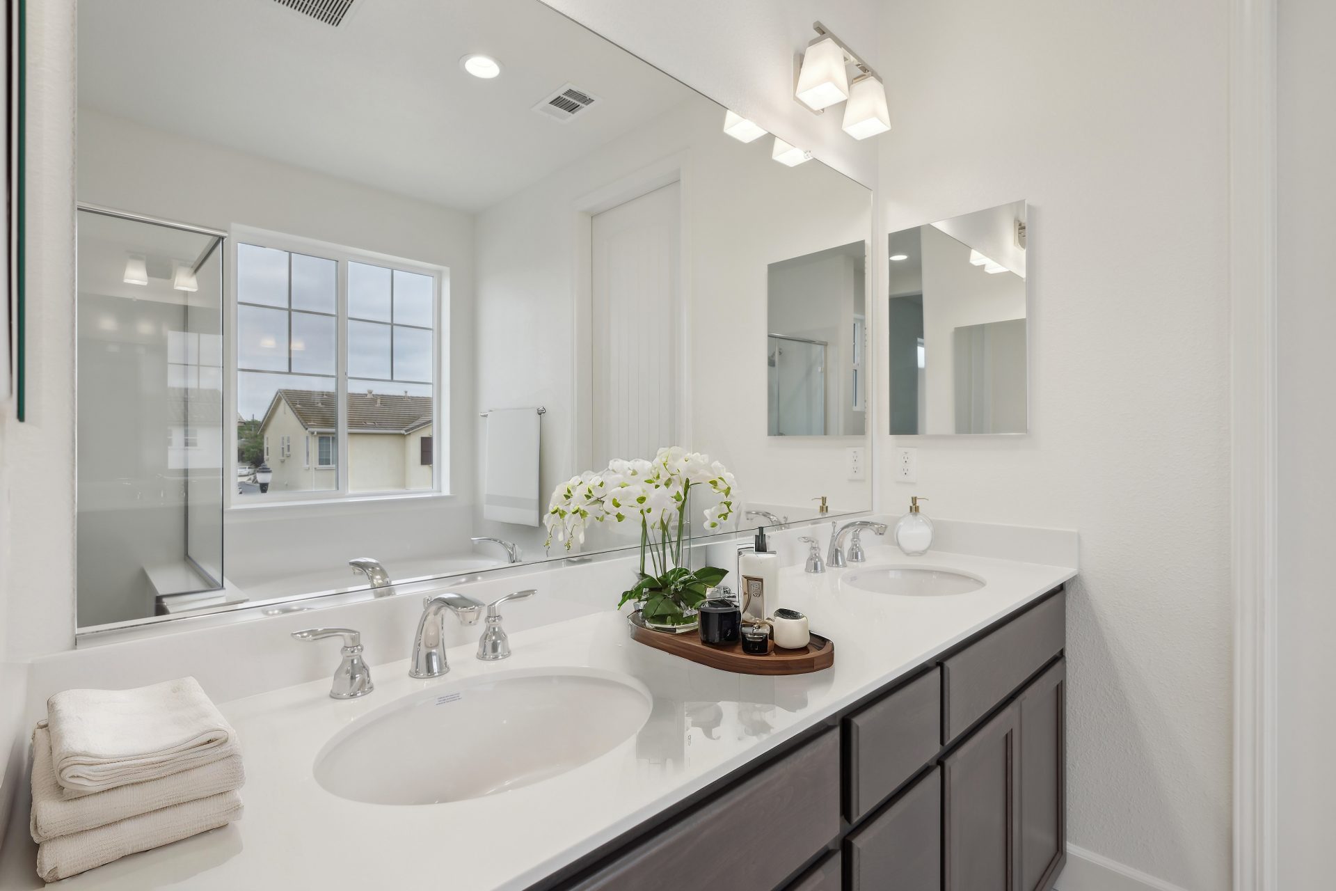 A bathroom with a double sink vanity, chrome fixtures, white countertop and brown stained cabinets. A long wall mirror with a light fixture above and décor items on the counter.