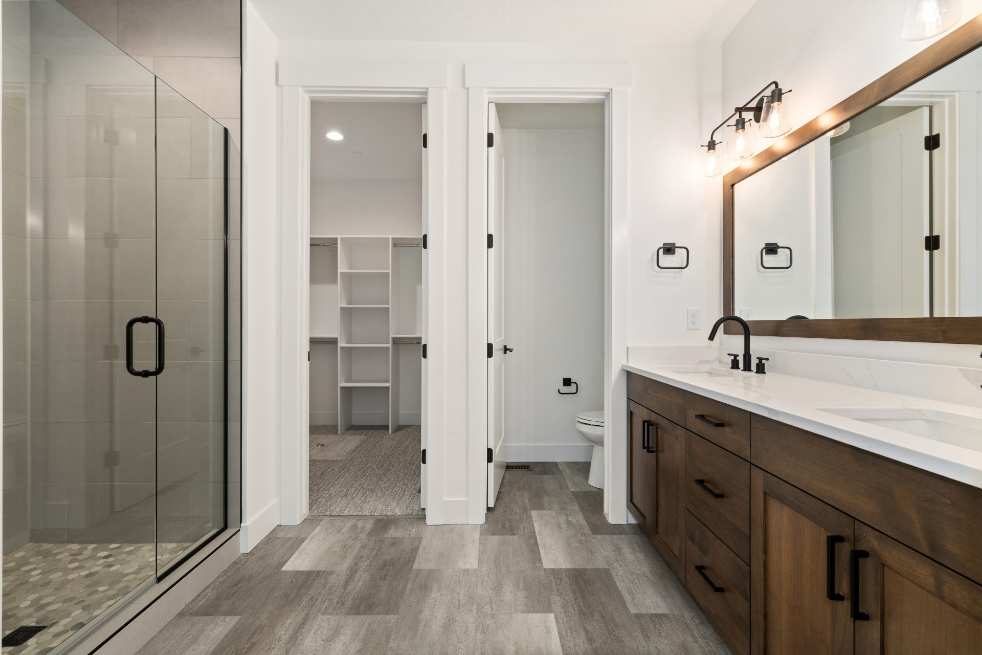 A bathroom with dual sink vanity, brown stained cabinetry and matching trim around the mirror. The faucets and light fixtures in a matte black finish with white countertops and medium brown floor tile.