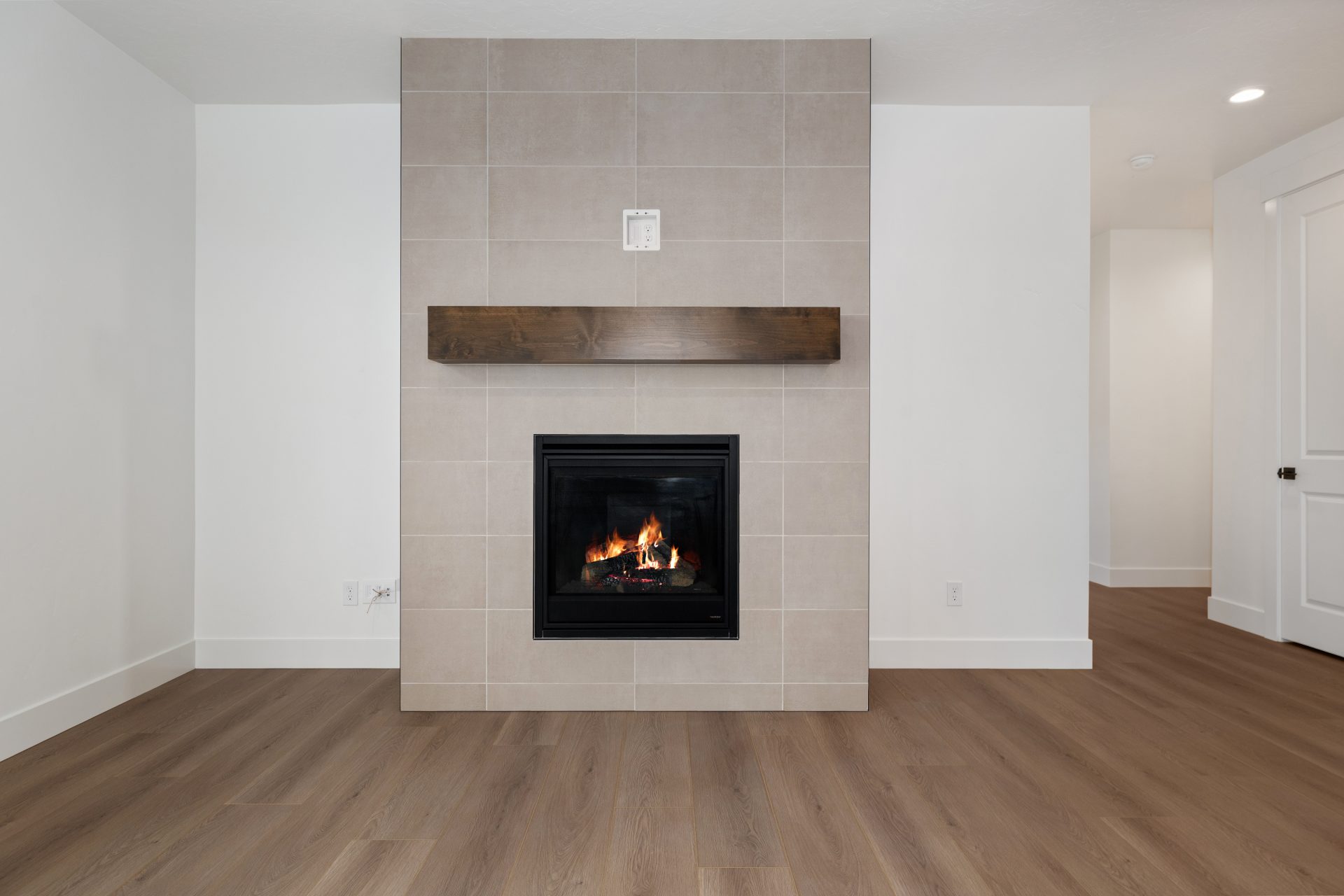 A fireplace with floor to ceiling tile detail and a wooden mantle, medium brown wood flooring and white color walls.
