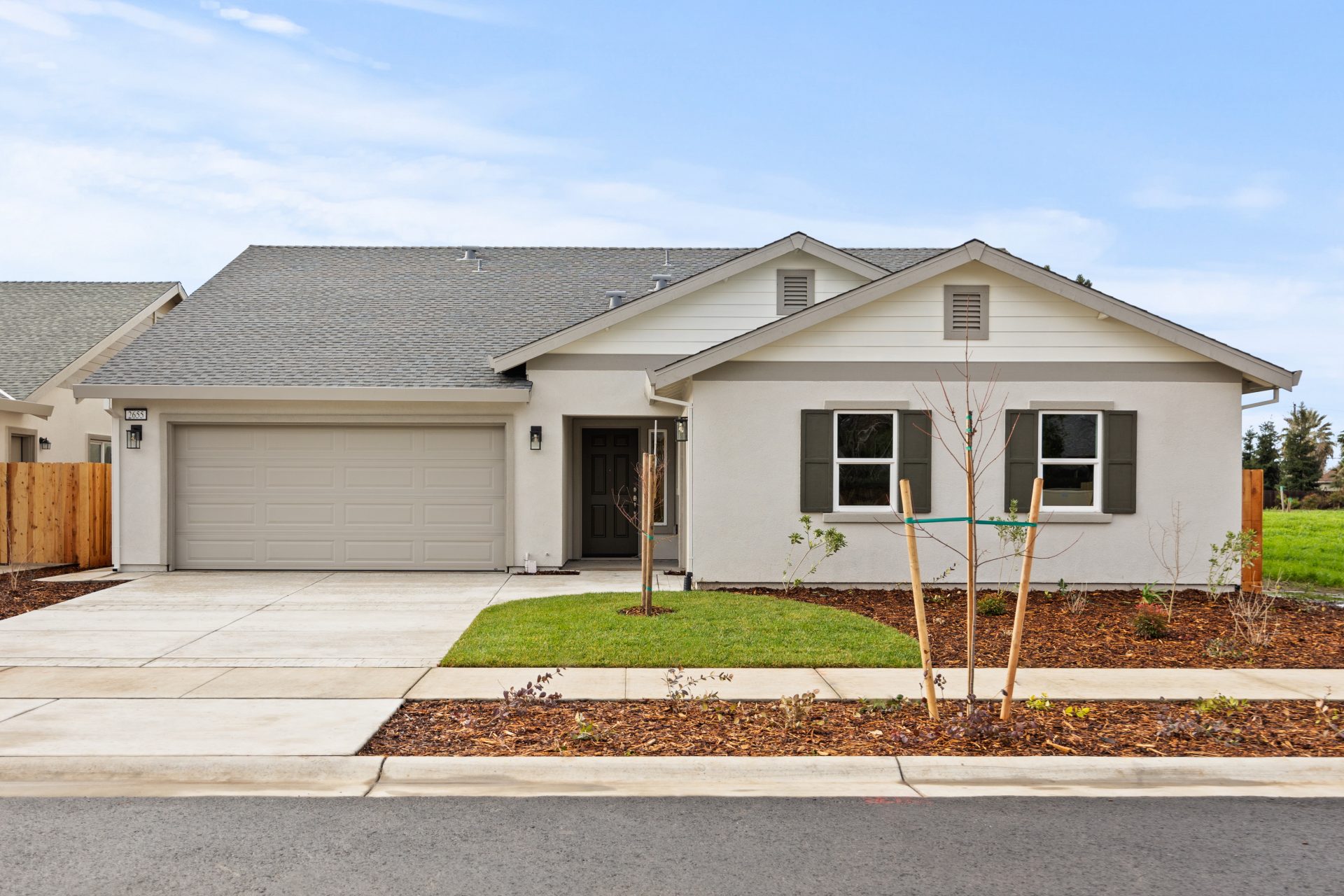 Exterior elevation of single-story home with light colored paint and grey trim. Home has mulch and grass landscaping with 2 young trees in front yard and blue sky in background