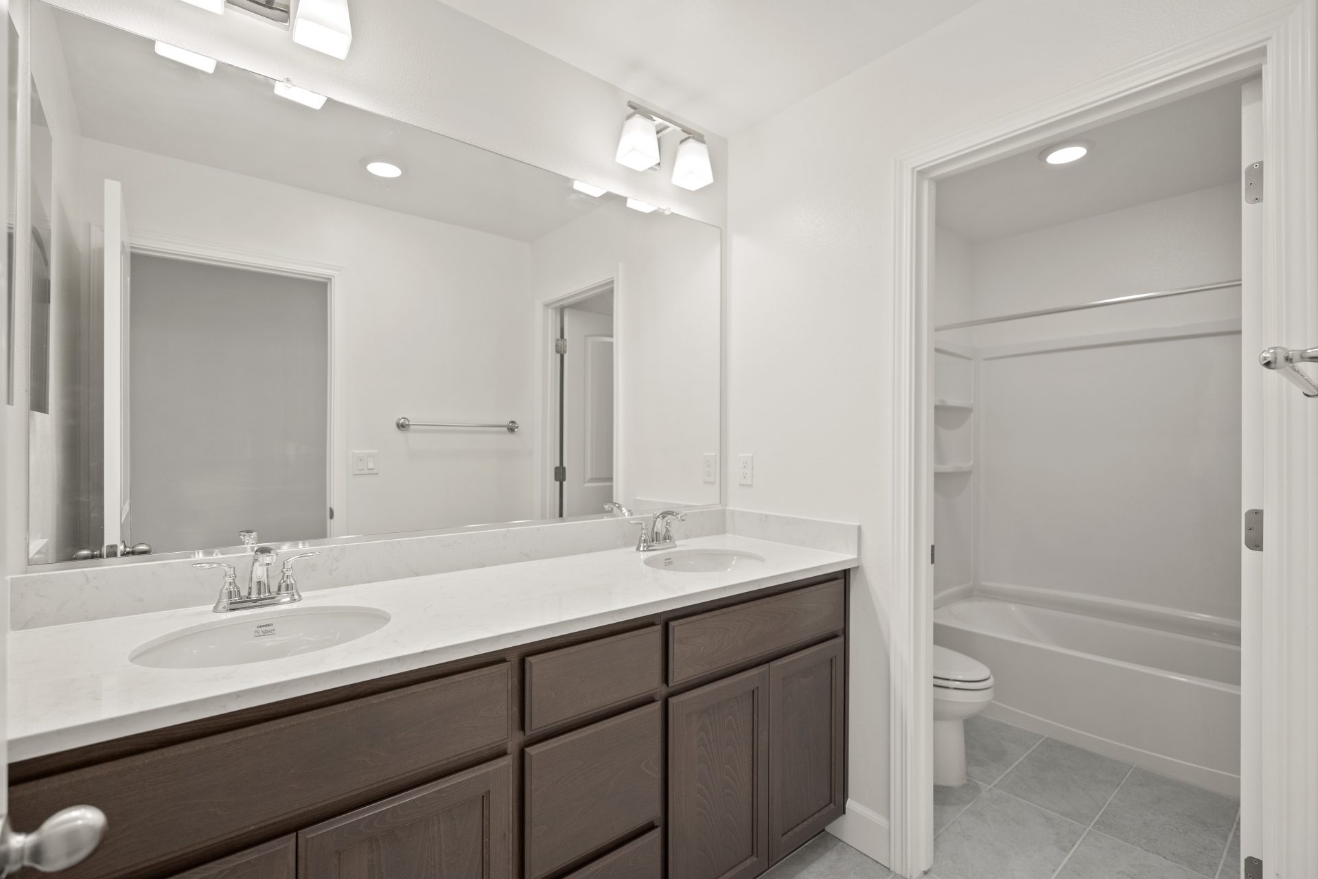 A two sink bathroom vanity with brown stained cabinets, and a light counter top. Light color tile flooring, chrome sink faucets, a long mirror and two light fixtures. View of the toilet area in the background