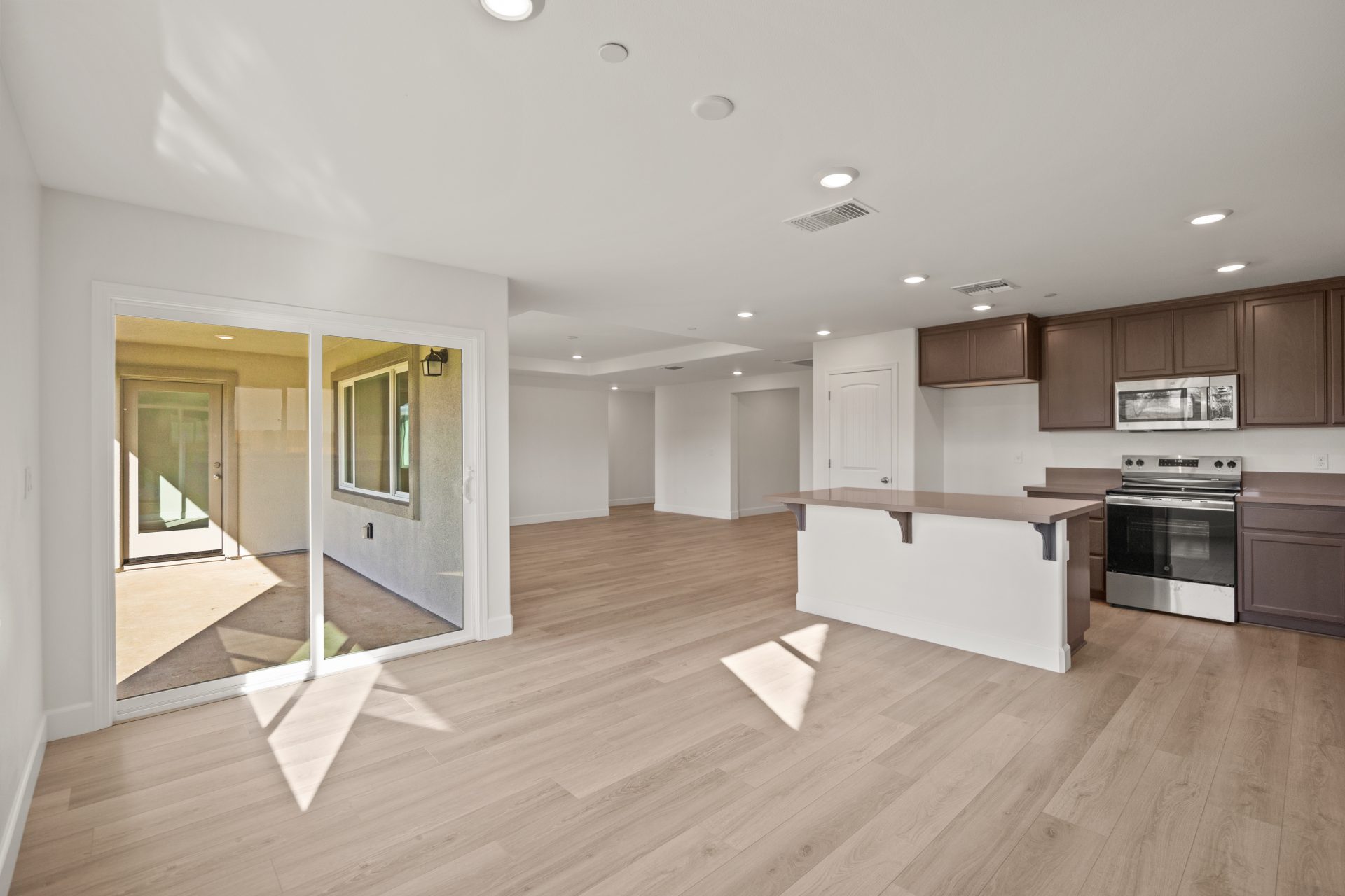 An unfurnished open space with light color wood floors. To the left a sliding glass door, to the right a kitchen with a kitchen island, light color walls and can recessed lighting in the ceiling.