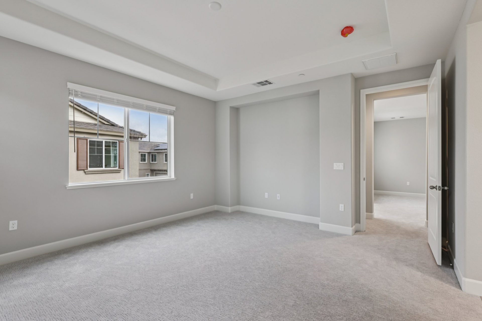A large unfurnished bedroom with light gray carpet, gray walls, white baseboards and ceiling, with an inset space in the ceiling. A window is on the left wall.
