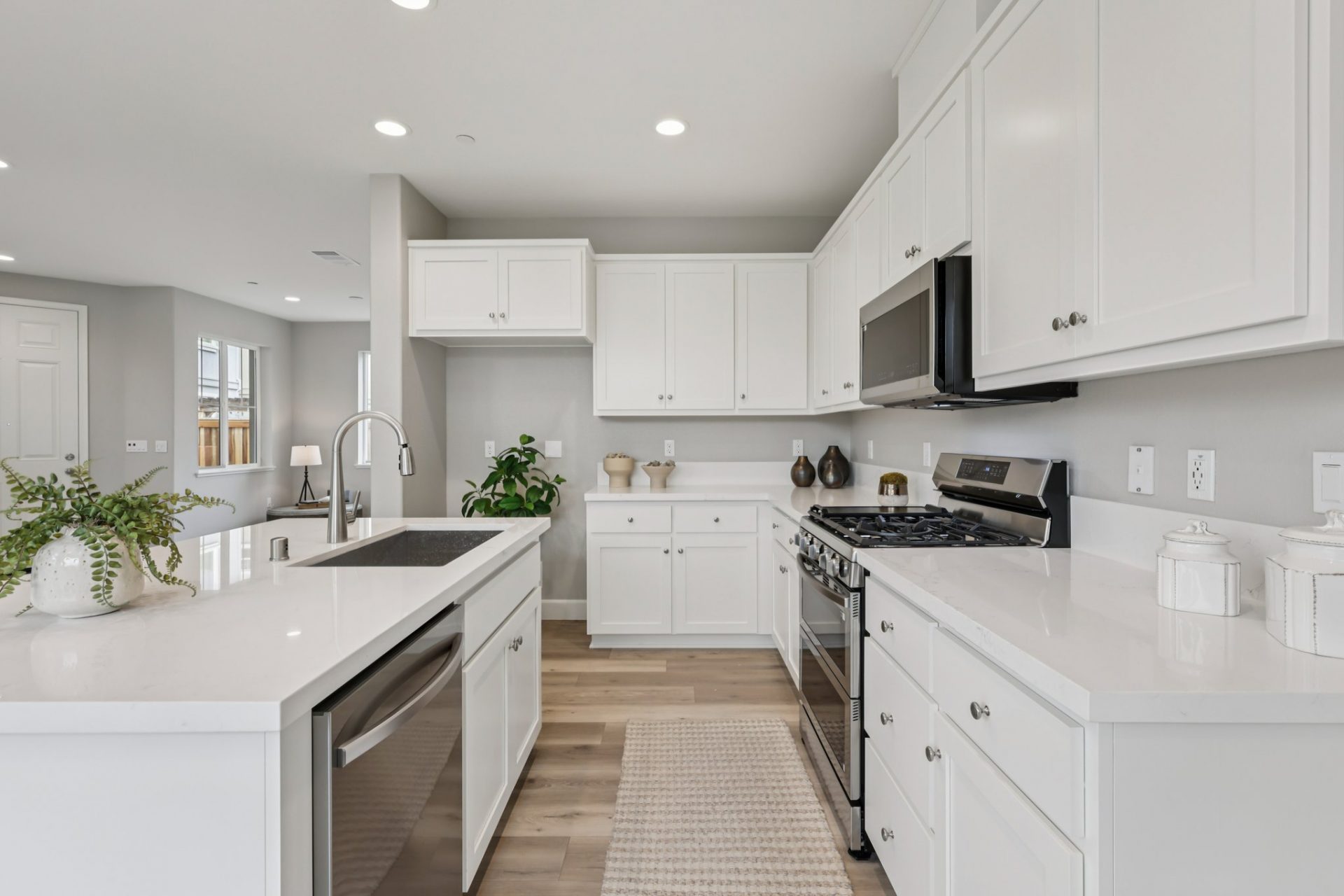 A kitchen with an island to the left with a sink and brushed nickel faucet, stainless steel dishwasher, upper and lower cabinets to the right with a stainless steel gas range stove with a microwave hood. Cabinets and countertops are white with nickel knob