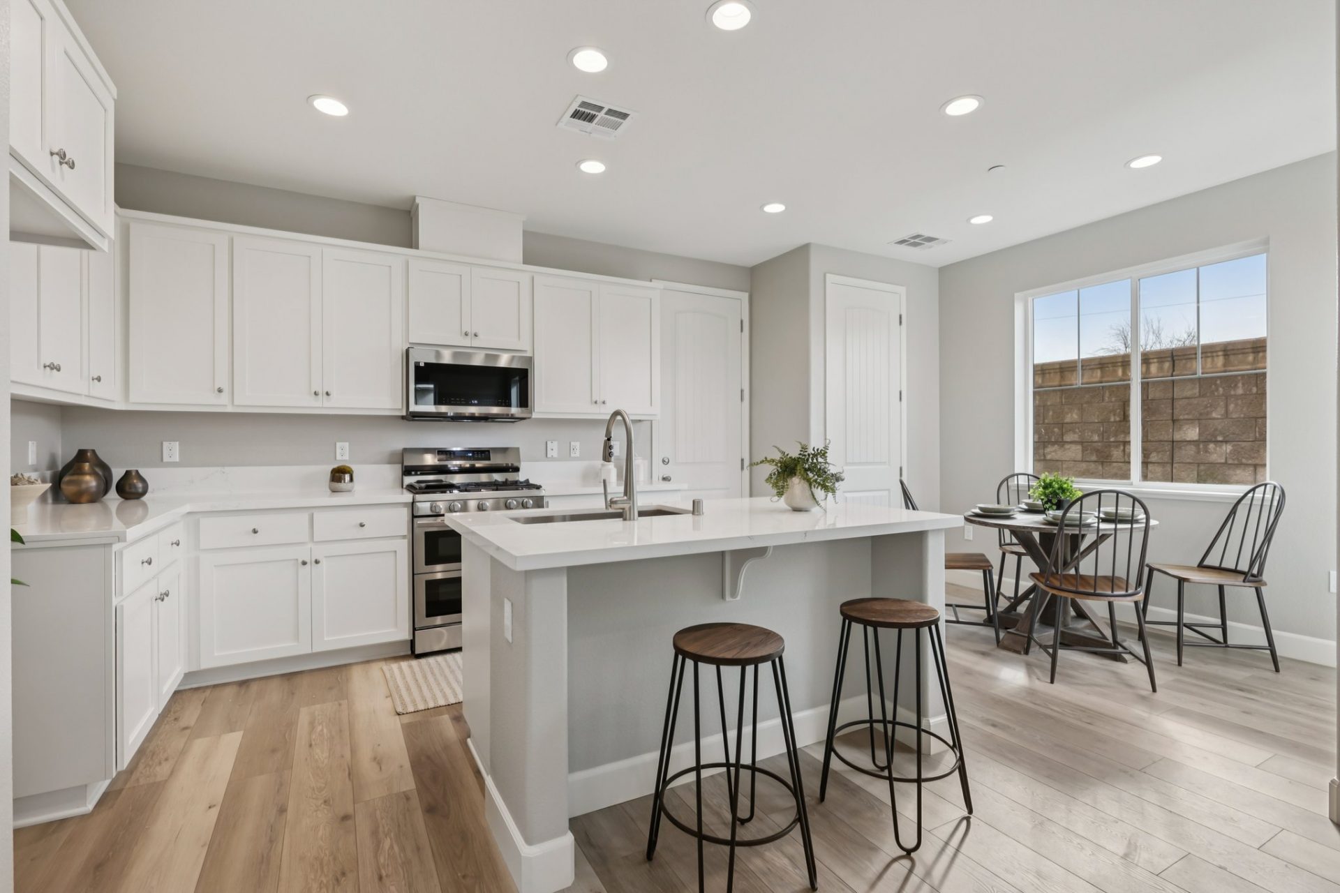 View of a kitchen with a dining nook to the right. The cabinets are white with nickel knobs, countertop is white with a brushed nickel faucet, stainless steel appliances and kitchen island. The flooring is of a light wood color and there are various decor