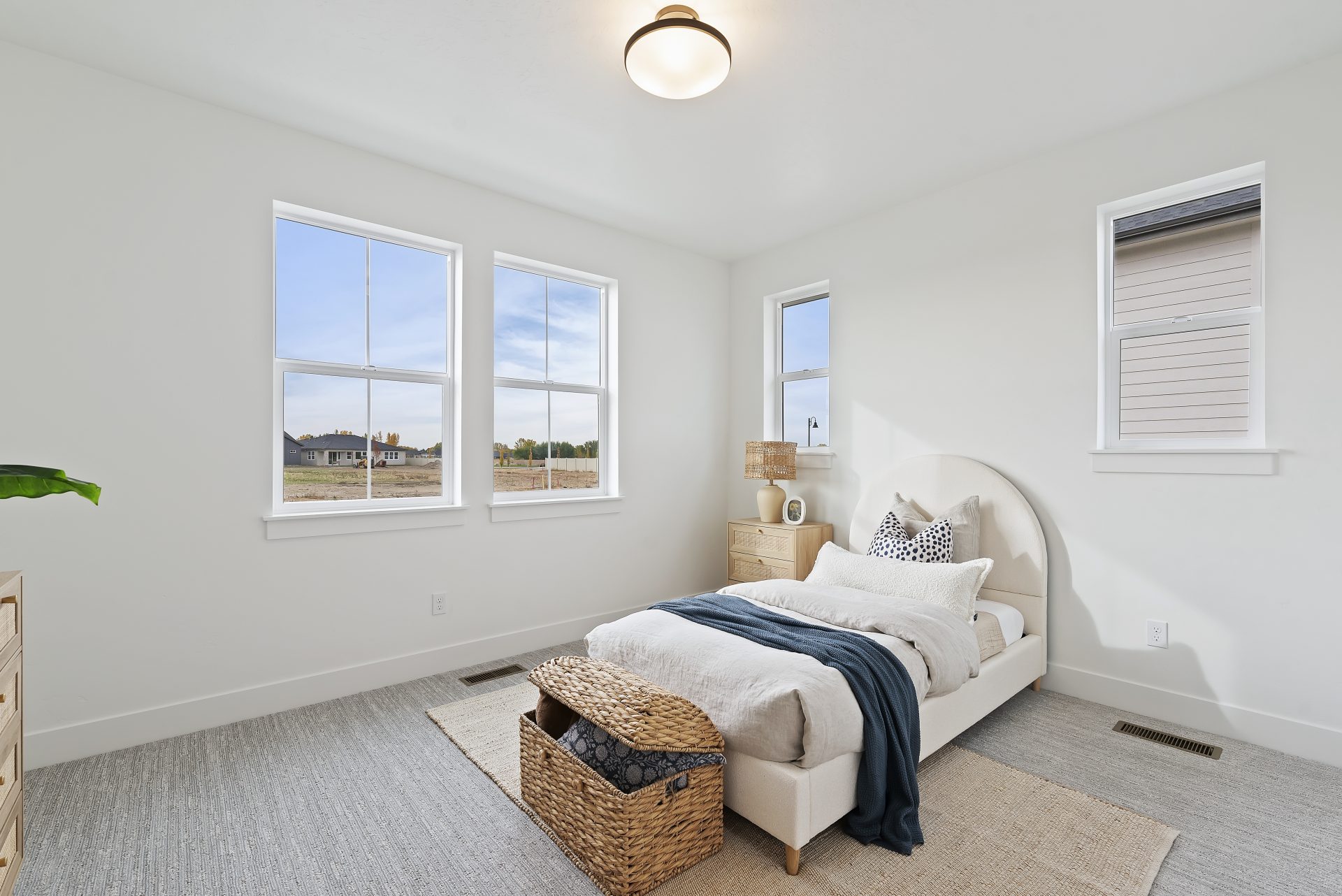A bedroom staged with a twin bed and furniture items, gray carpet light color walls, four windows and a light fixture in the ceiling.