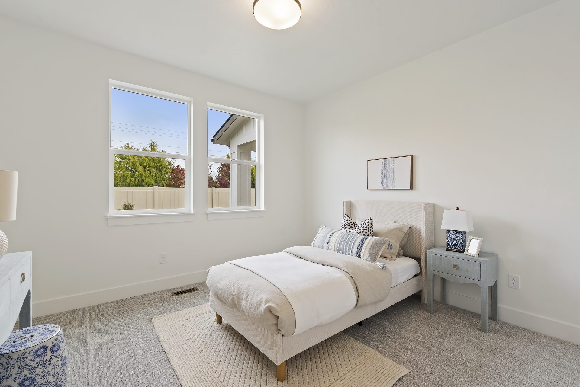 A bedroom staged with a twin bed and furniture items, gray carpet light color walls, two windows and a light fixture in the ceiling.