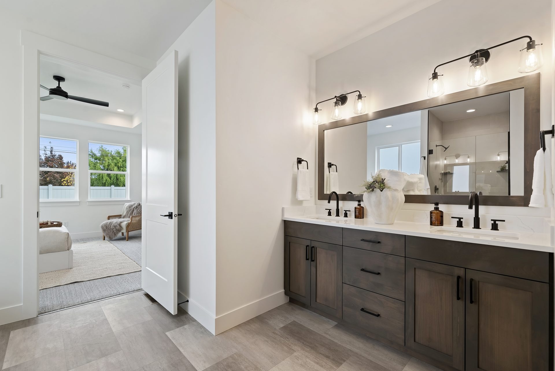 A bathroom with tile flooring a dual sink vanity, with brown stain grade cabinets, black pulls and black faucets, white countertops with a 6" backsplash, a framed mirror spanning the length of the vanity and two- three light fixtures located above.