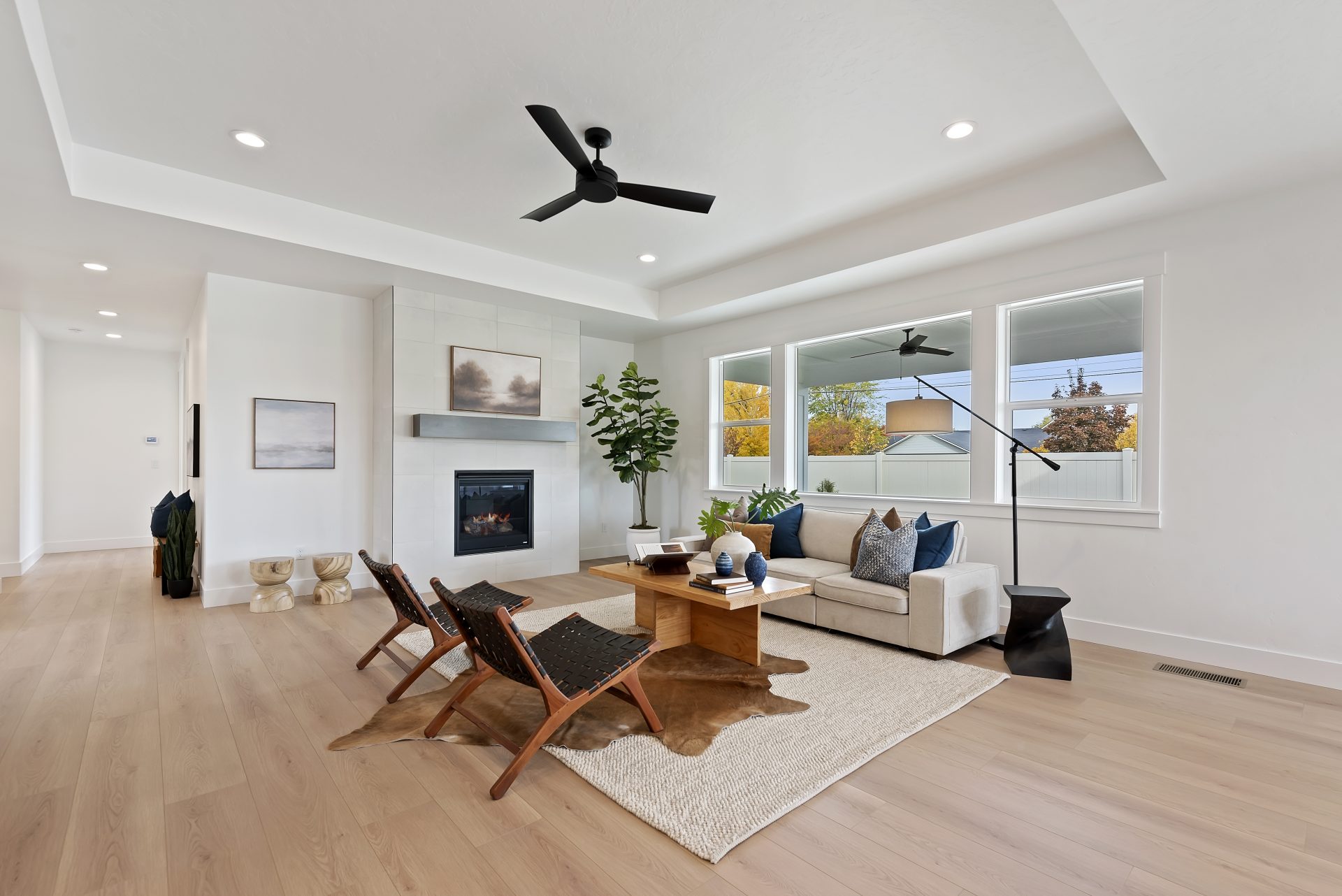 A staged living room with a couch, chairs and coffee table. Light brown wood floors, a gas fireplace with floor to ceiling tile and a mantle, can LED lighting in the ceiling and a ceiling fan over the seating area. Three windows create a bright space.